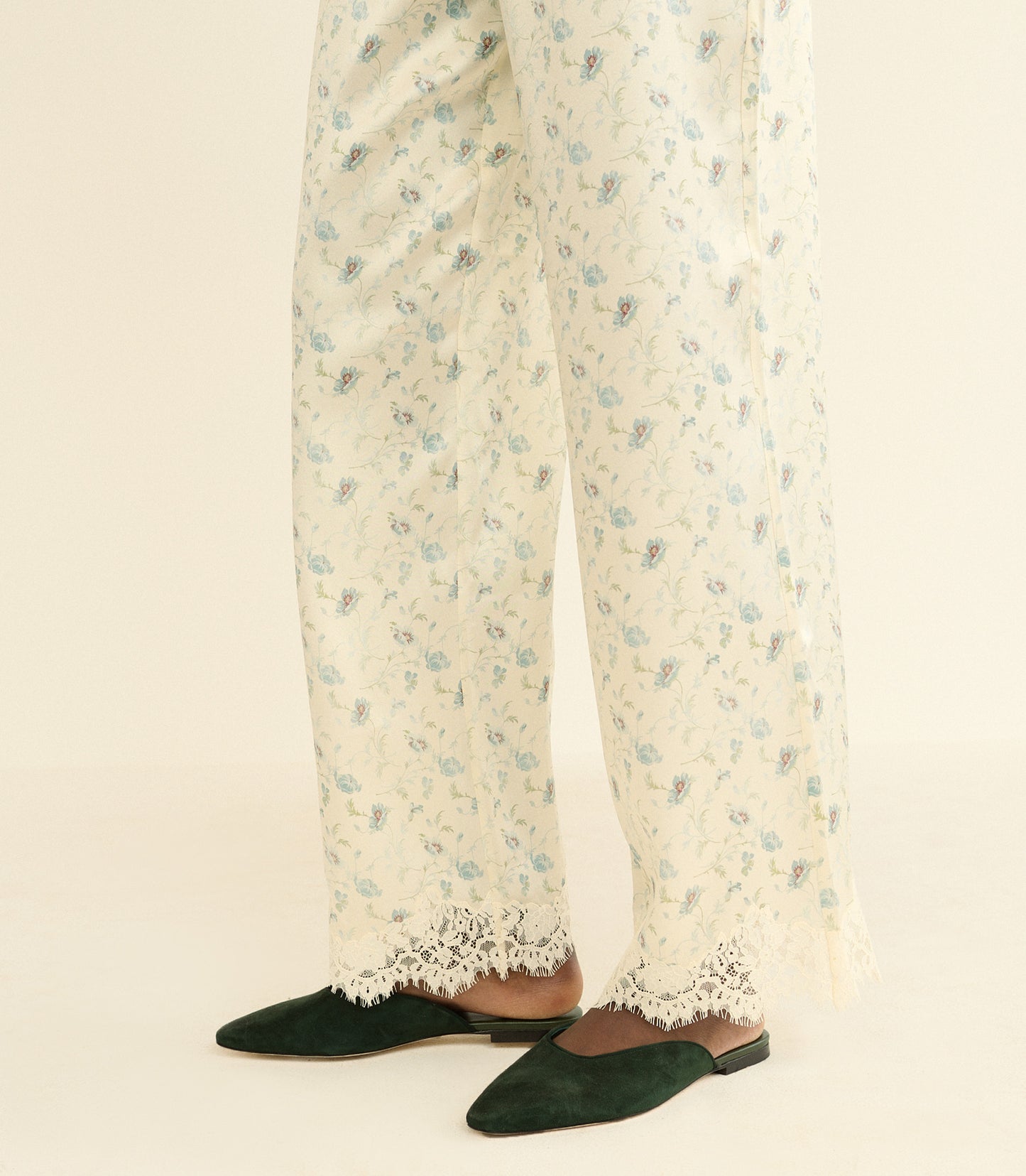 A close-up of a person's legs wearing cream-colored wide-leg trousers with a delicate blue and green floral print and lace trim, paired with dark green pointed-toe flats on a light beige background.