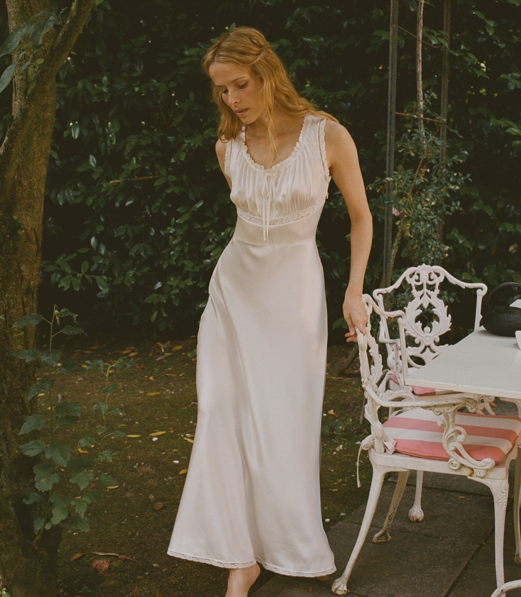 A woman wearing a sleeveless white satin maxi dress with lace detailing at the neckline, standing in a garden next to a white metal patio table and chair.