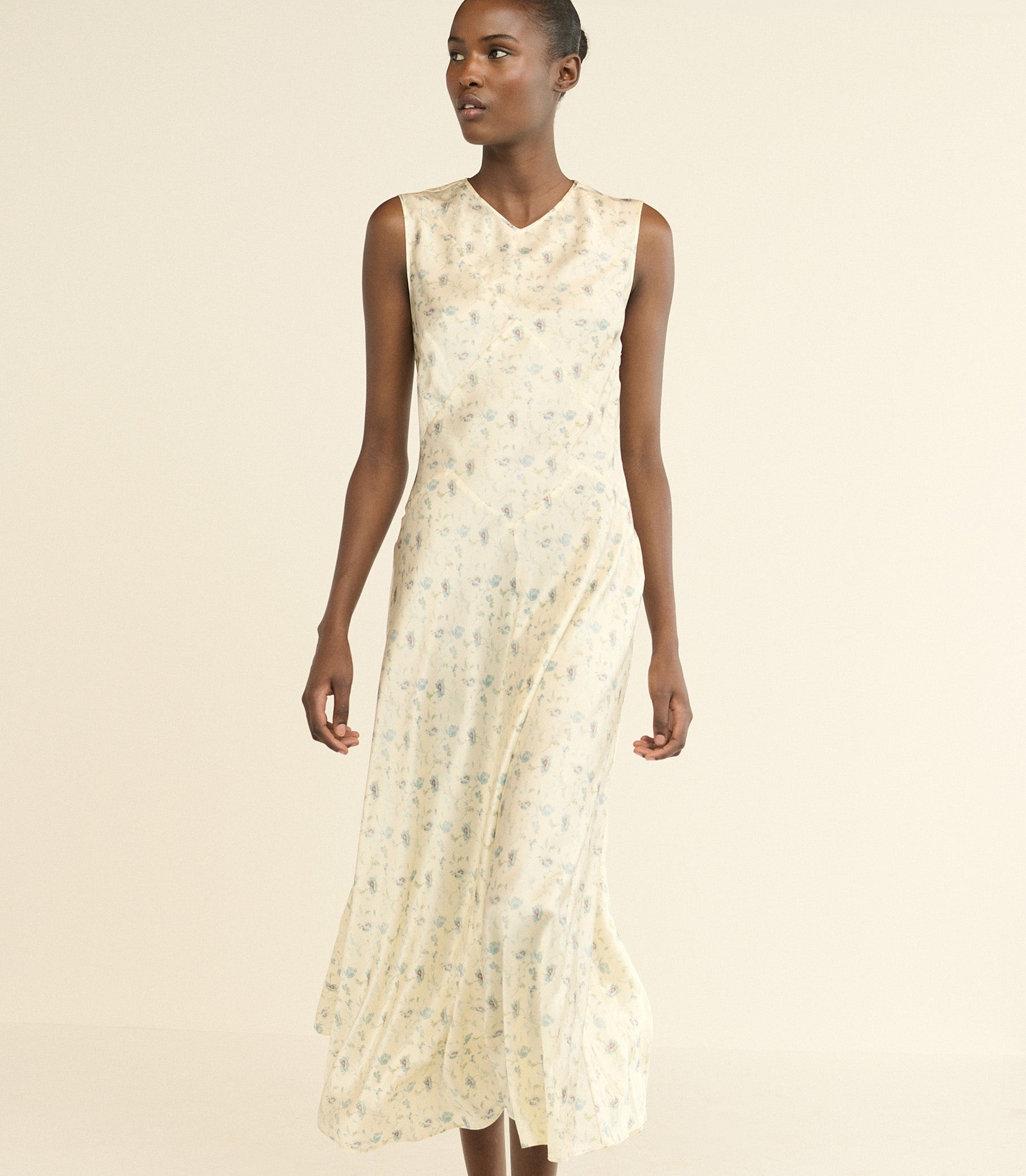 A sleeveless, V-neck midi dress in a cream-colored silk fabric with a delicate floral print, modeled against a solid light beige background. 1
