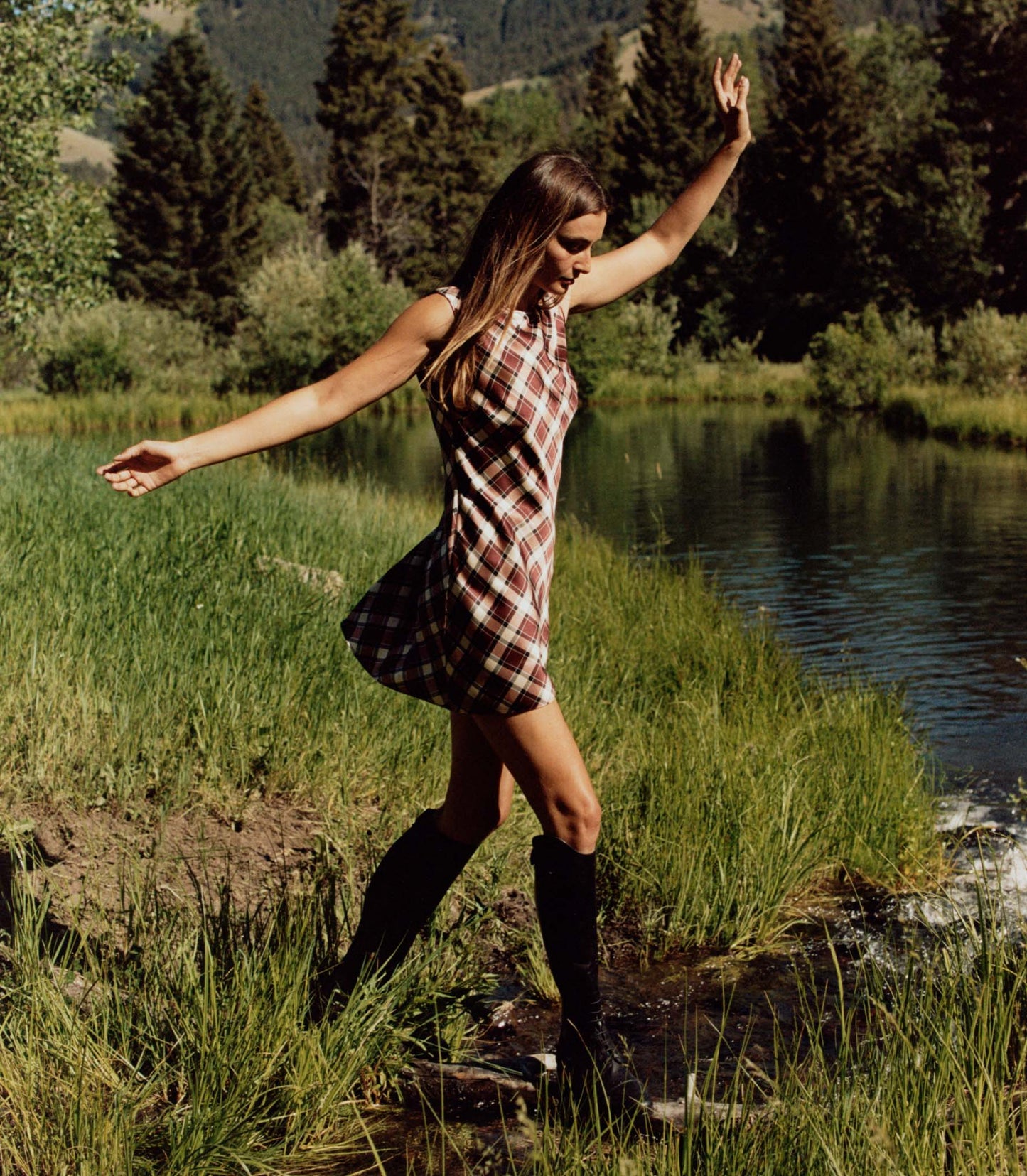 Woman in a plaid dress and knee-high boots standing by a lake with arms outstretched.