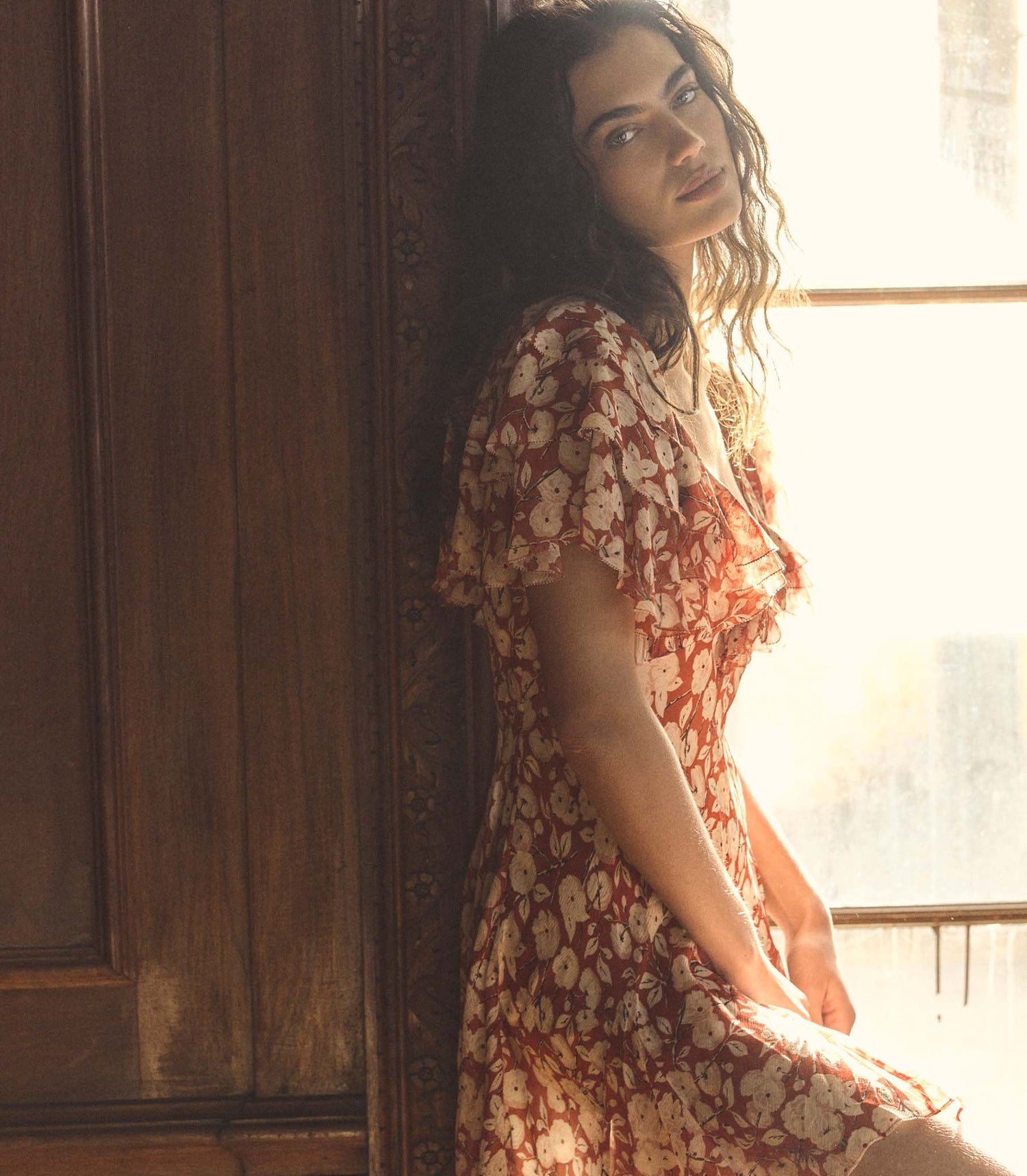 Woman in a floral dress standing by a wooden door with sunlight streaming in.