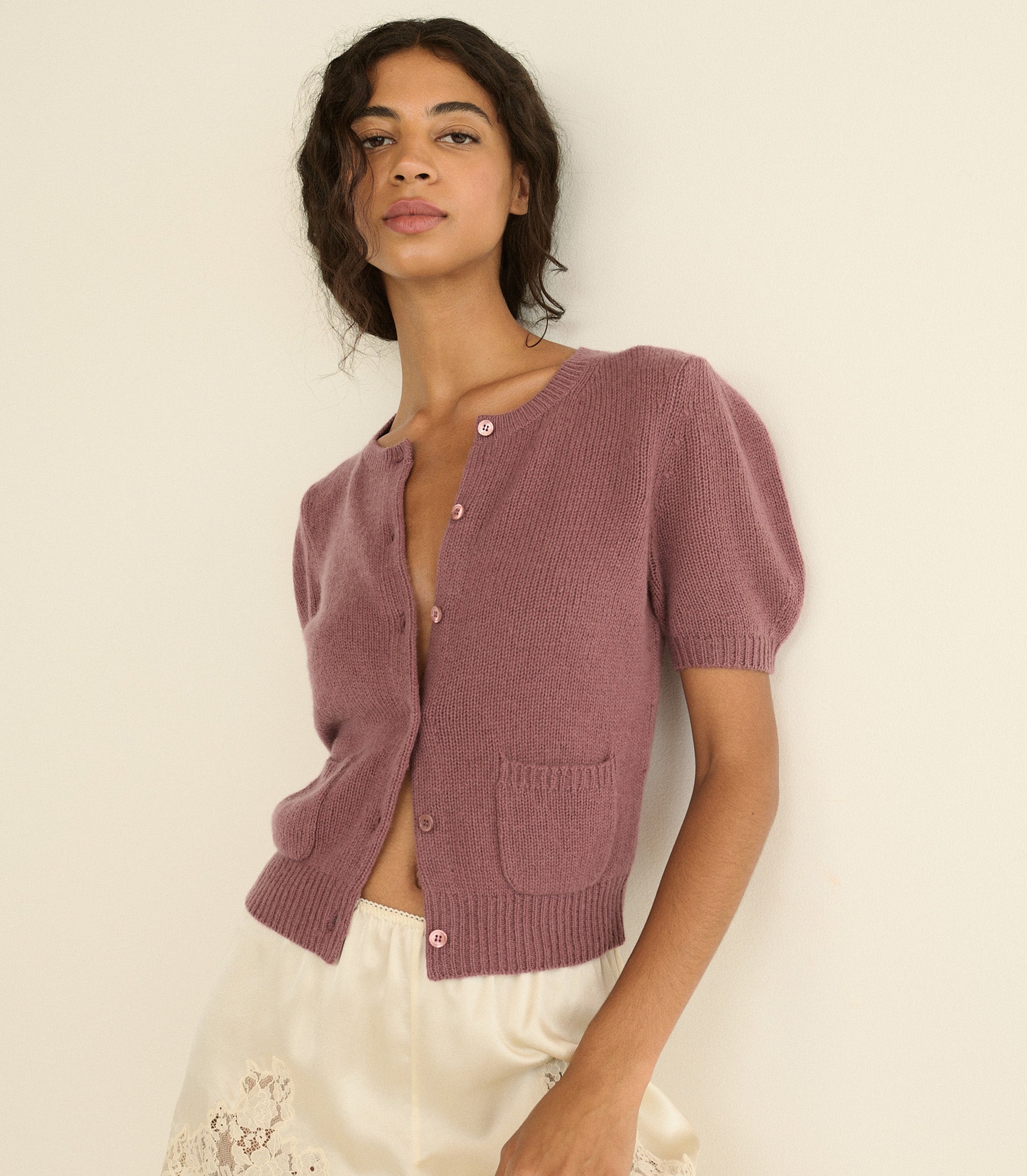 A woman wearing a short-sleeve knit cardigan in a dusty mauve-pink color, paired with white-colored shorts, posing against a plain off-white background. 1
