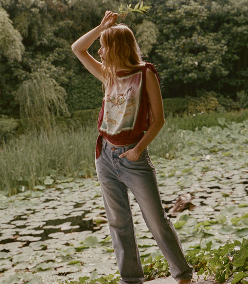 A woman stands outdoors in a garden pond wearing a sleeveless graphic top with a red draped layer, light gray jeans, and bare feet. The background features green foliage and lily pads.
