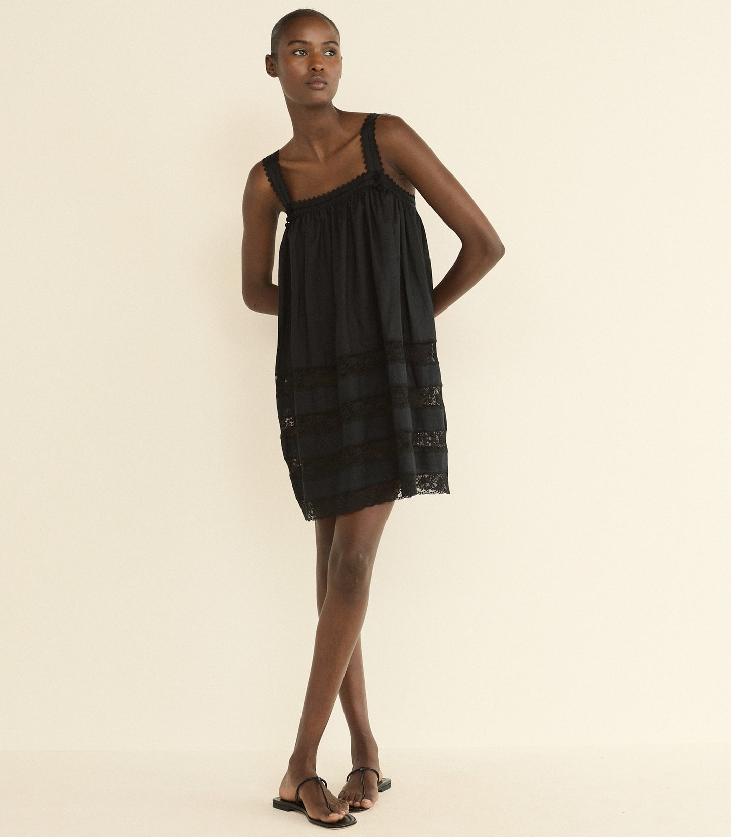 A model wearing a black sleeveless mini dress with a gathered bodice and sheer lace-like horizontal bands near the hem, paired with black sandals, against a plain cream background.