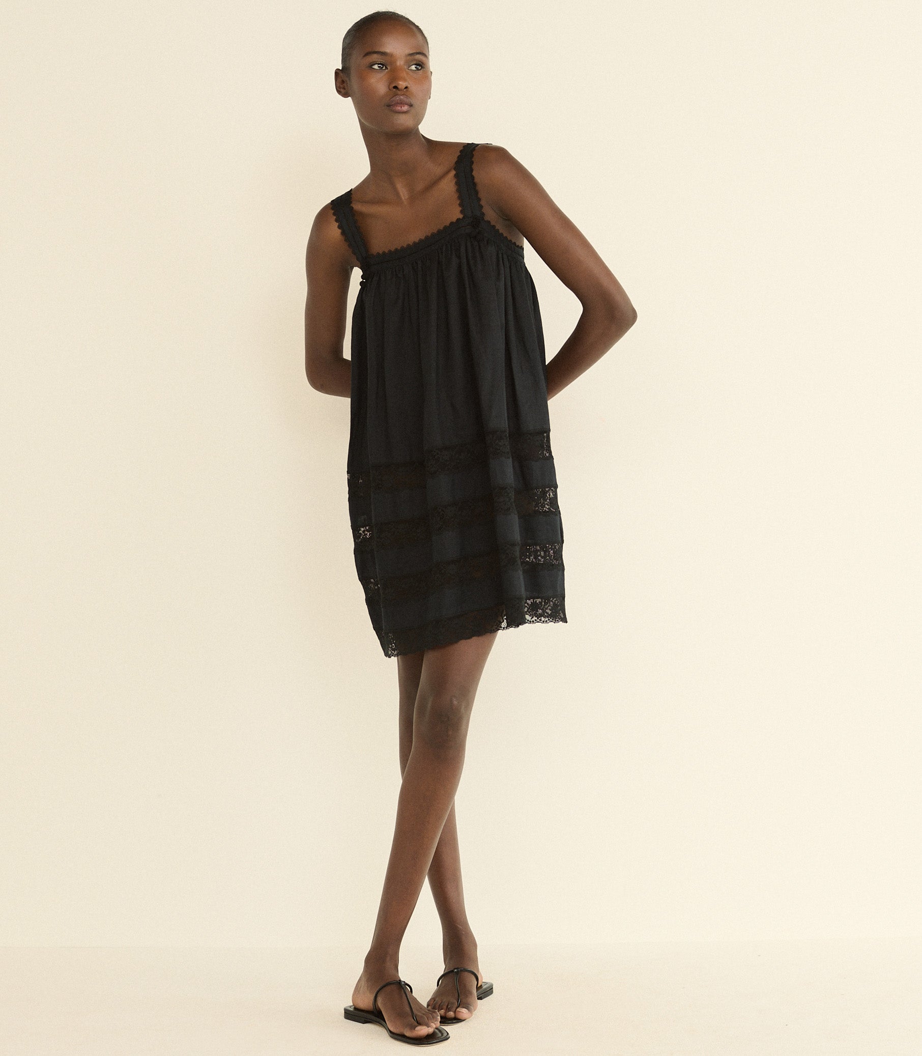 A model wearing a black sleeveless mini dress with a gathered bodice and sheer lace-like horizontal bands near the hem, paired with black sandals, against a plain cream background.
