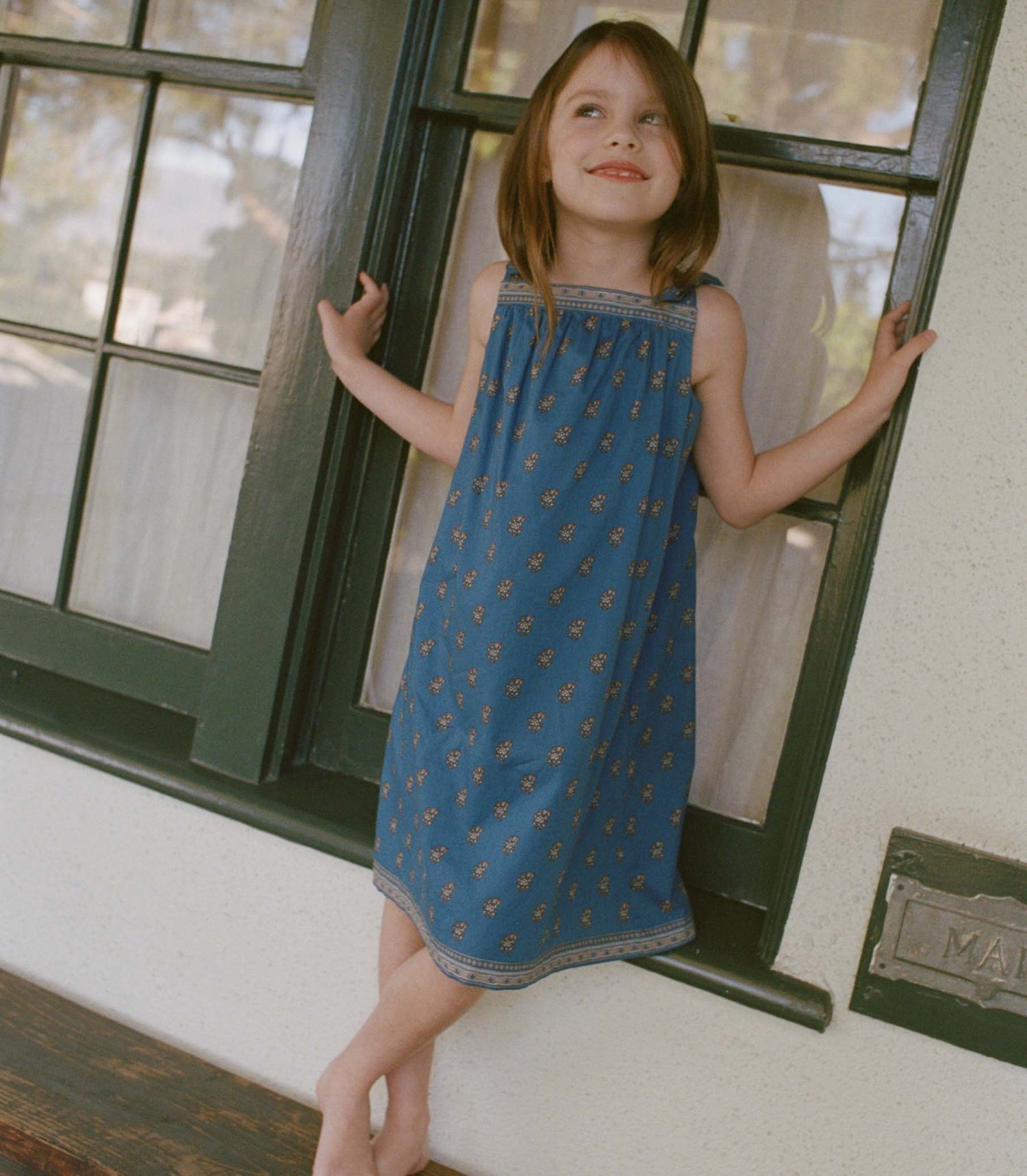 Young girl in a blue paisley dress standing by a window