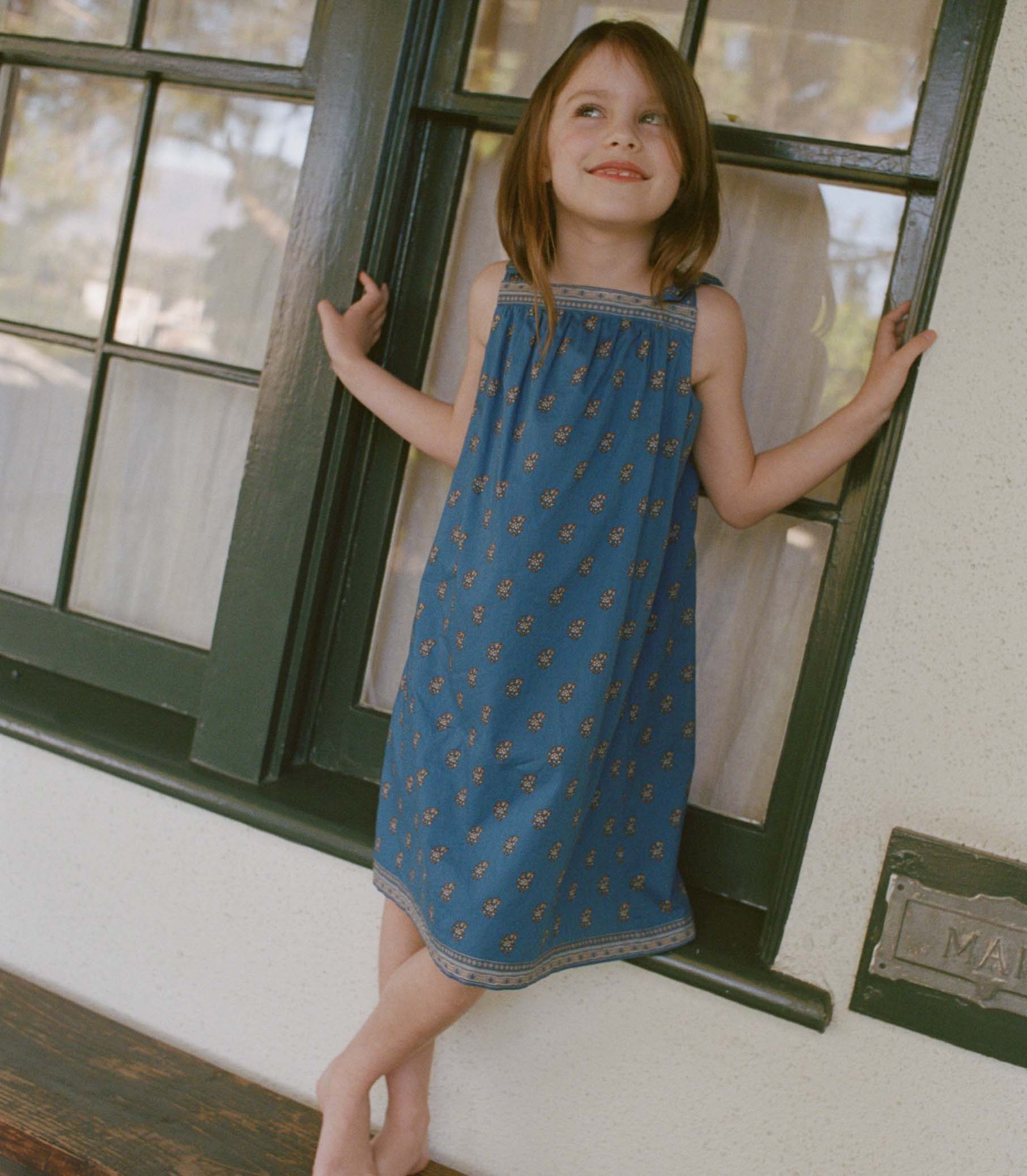 Young girl in a blue paisley dress standing by a window