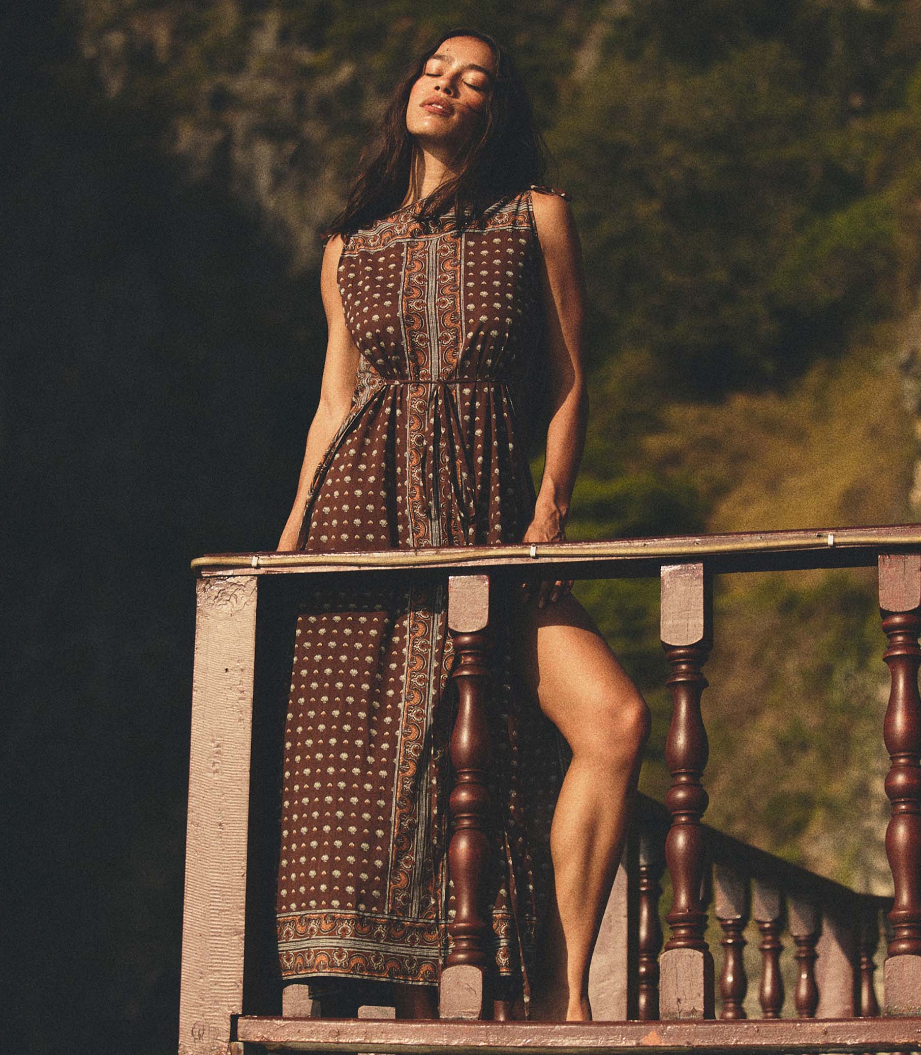 Woman in a patterned dress standing on a wooden railing with a blurred natural background 1
