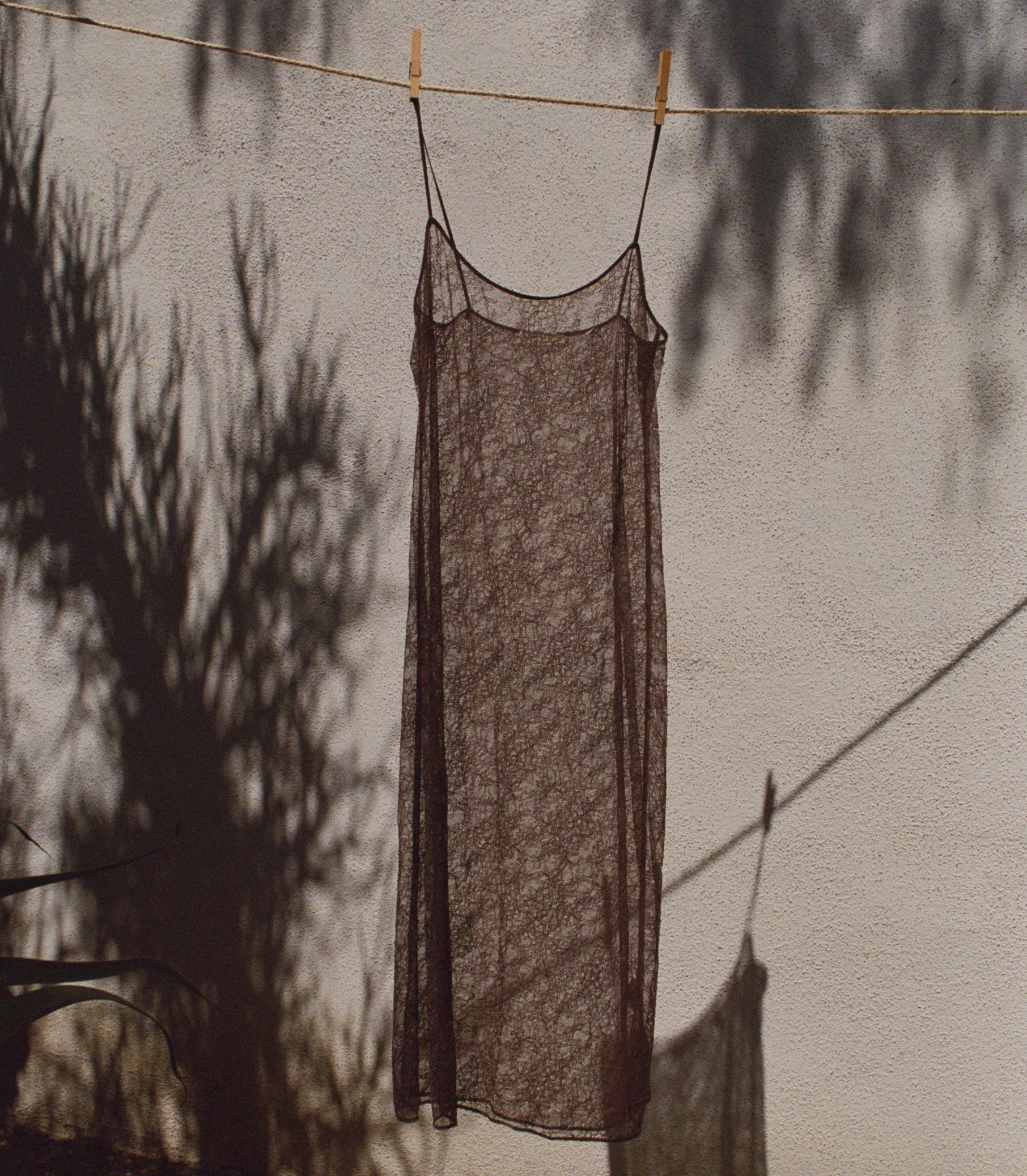 A sheer, dark brown lace slip dress hanging on a clothesline against a textured wall with tree shadows cast across it. 1
