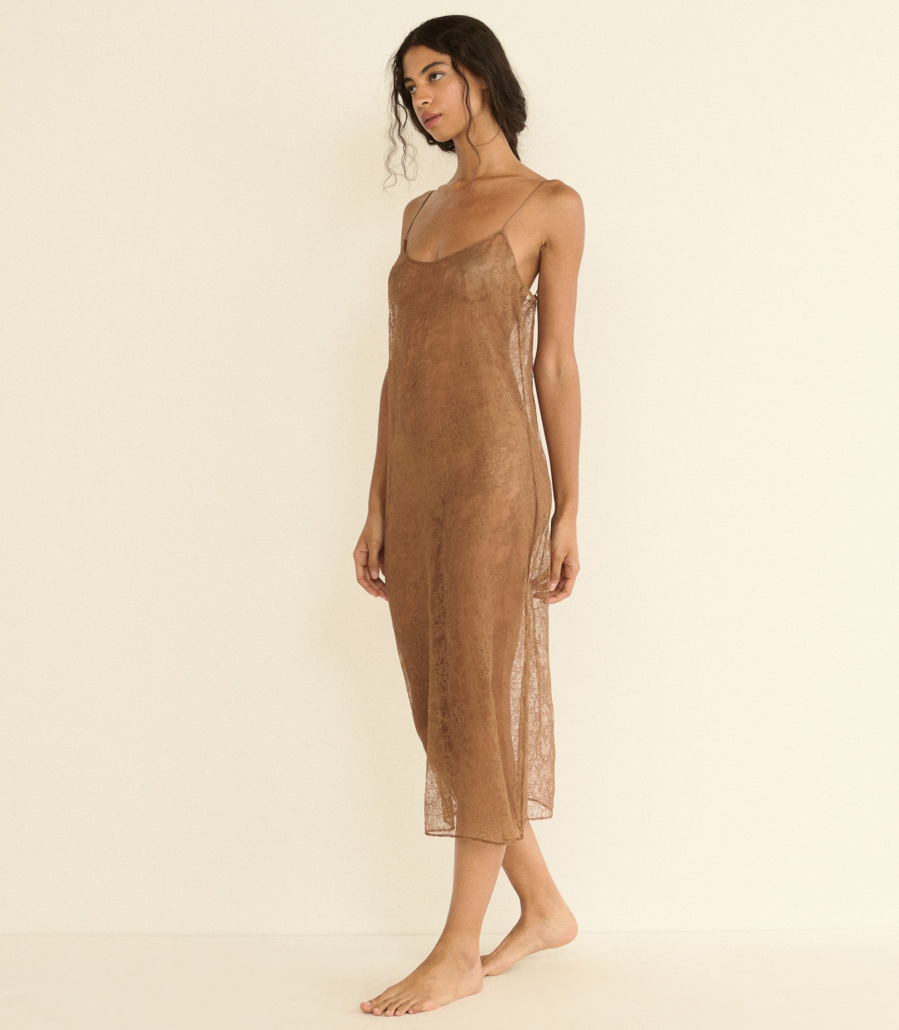 A woman modeling a sheer, lace brown slip dress with spaghetti straps against a plain, warm off-white studio background.