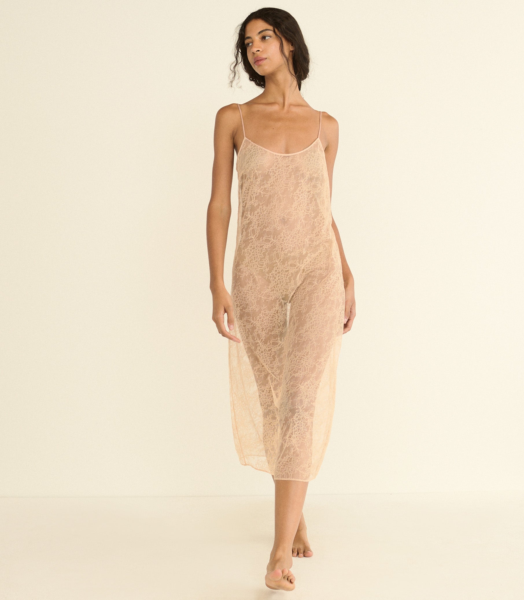 A woman modeling a sheer, nude-beige lace slip dress with thin spaghetti straps against a solid, warm off-white studio background.