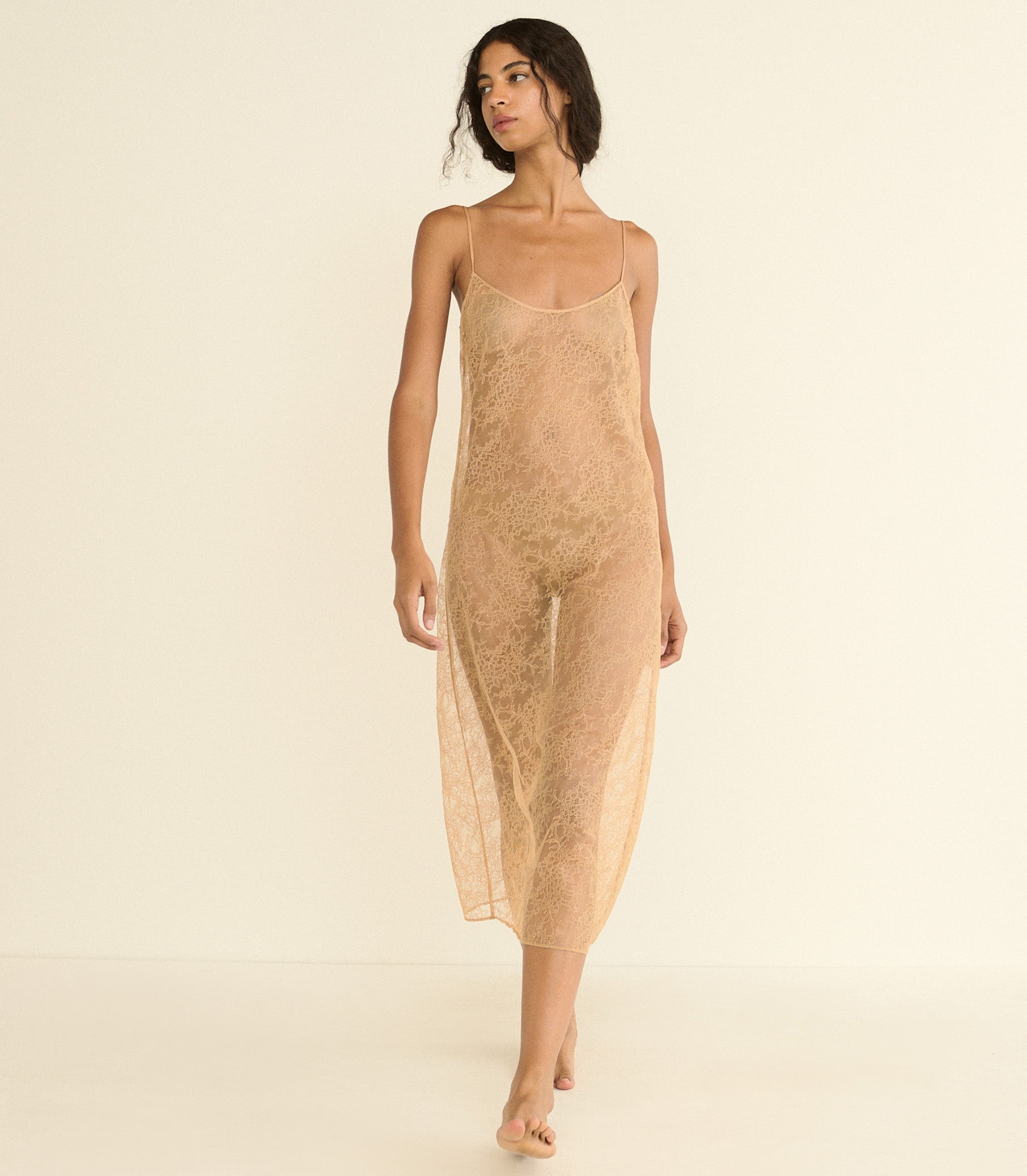 A woman modeling a sheer, textured slip dress in a light beige or tan color, standing against a solid, warm off-white studio background. 1
