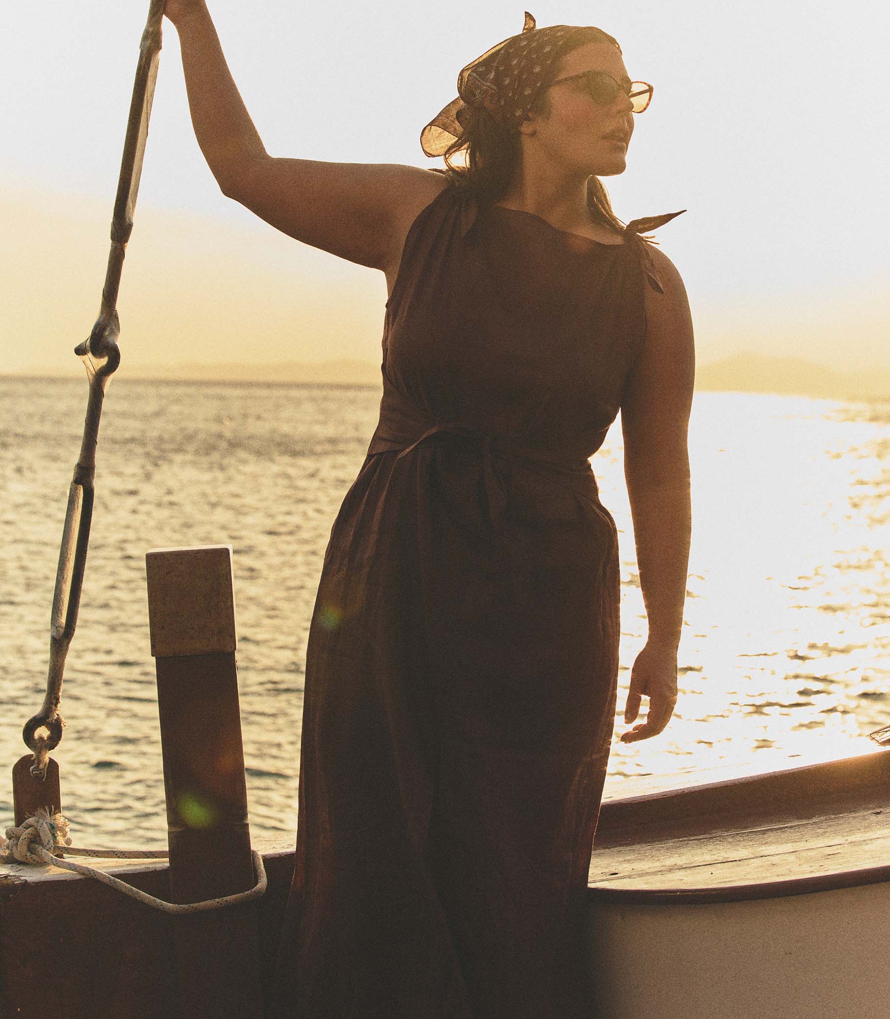 Woman in a brown dress standing on a boat with a sunset or sunrise in the background. 5
