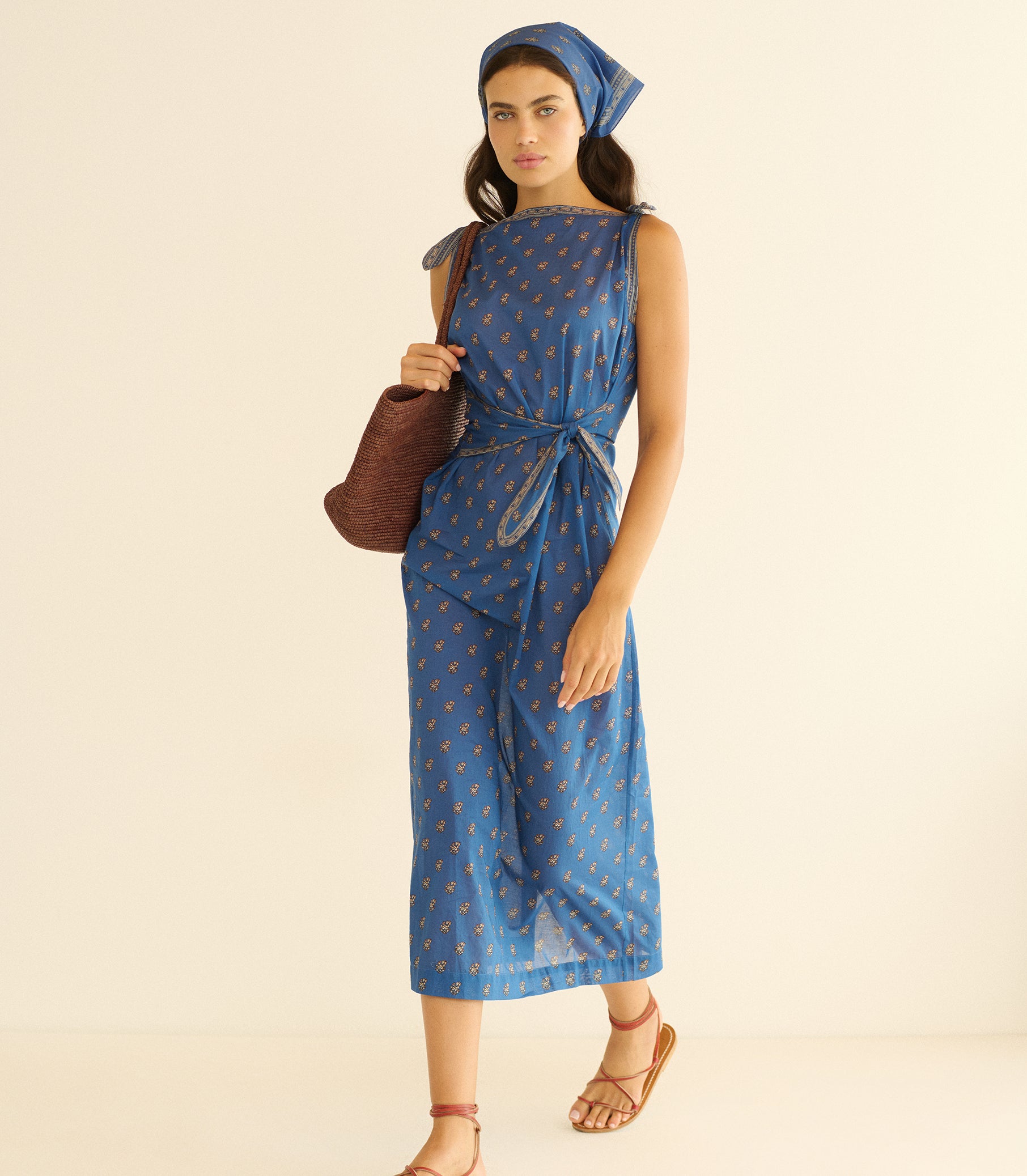 Woman wearing a blue paisley dress, holding a brown bag on a beige background 1
