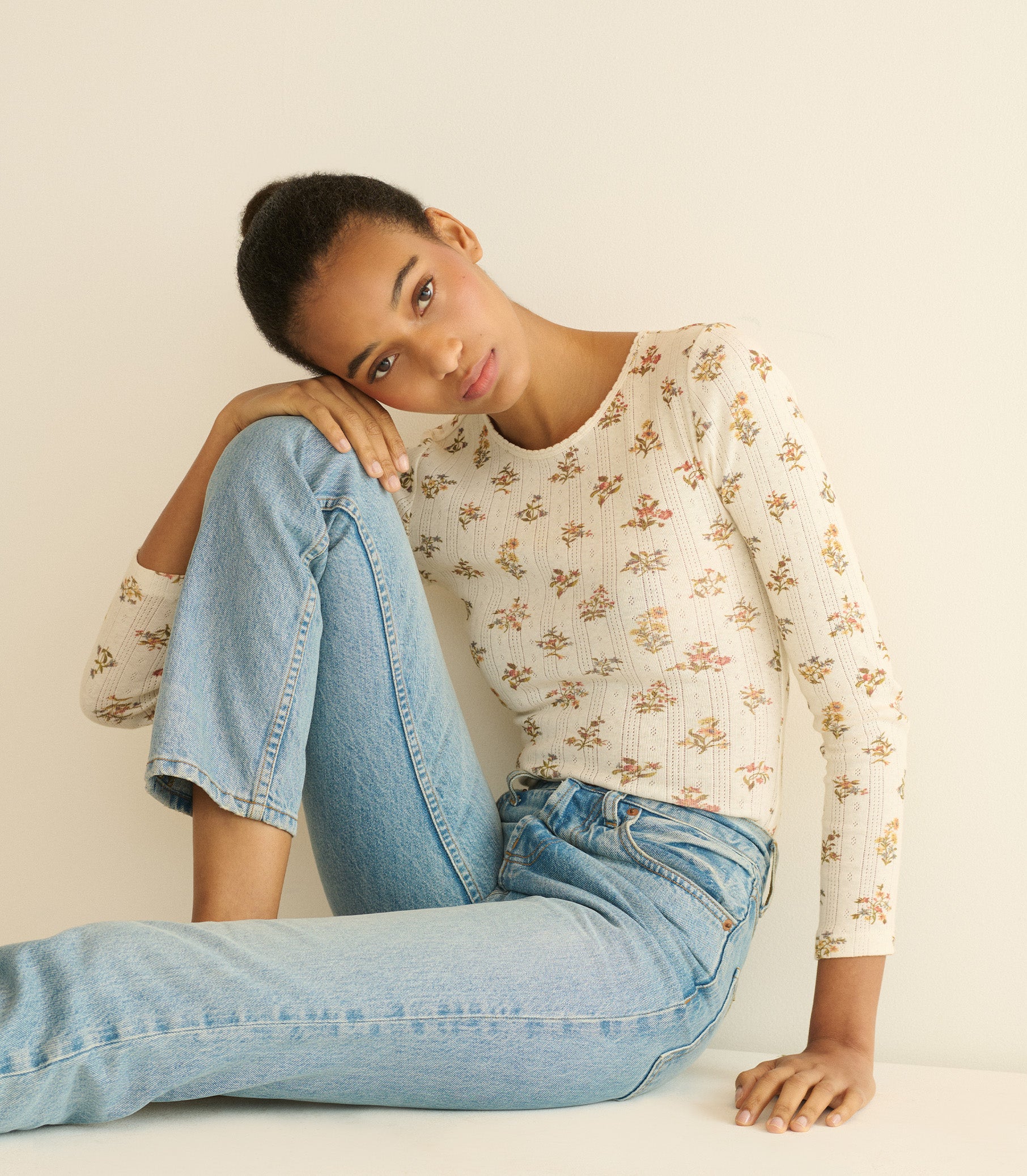 Woman wearing a floral top and blue jeans sitting on a white surface. 4
