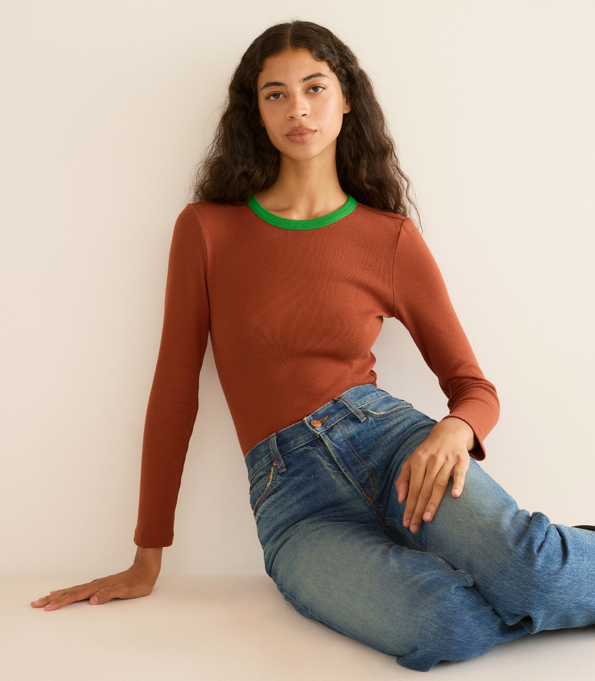 Woman wearing a brown sweater with green trim and blue jeans sitting against a white wall. 1
