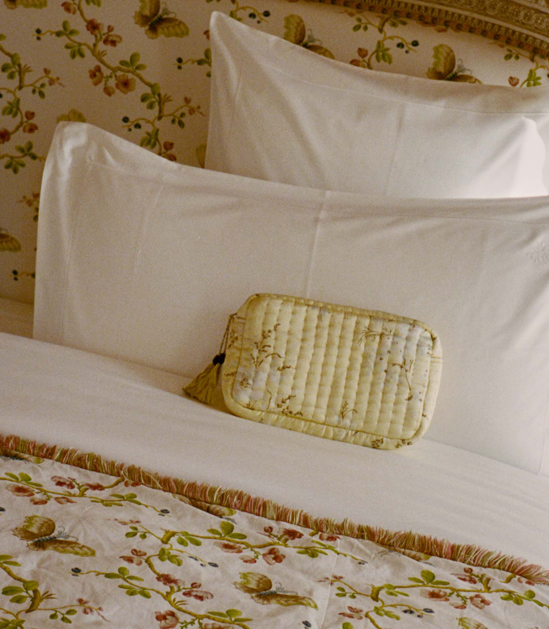 Light yellow floral pouch against white pillows on a bed with floral bedding and wallpaper. 1
