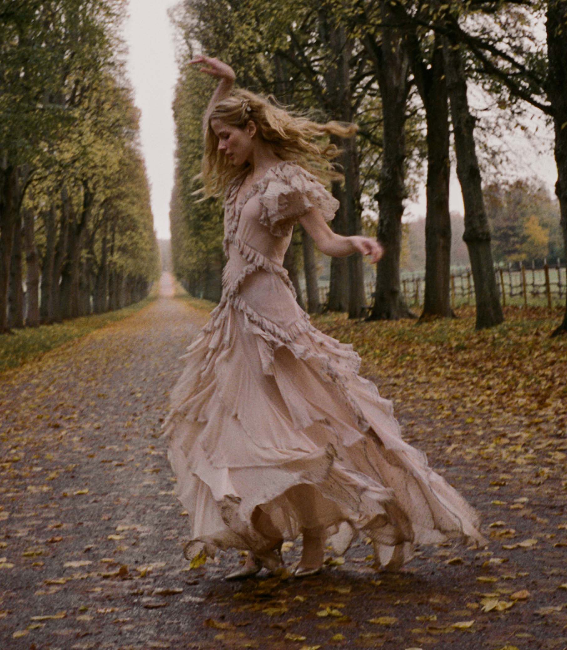 Woman in a long, antique blush dress dancing on a tree-lined path with fallen leaves. 1
