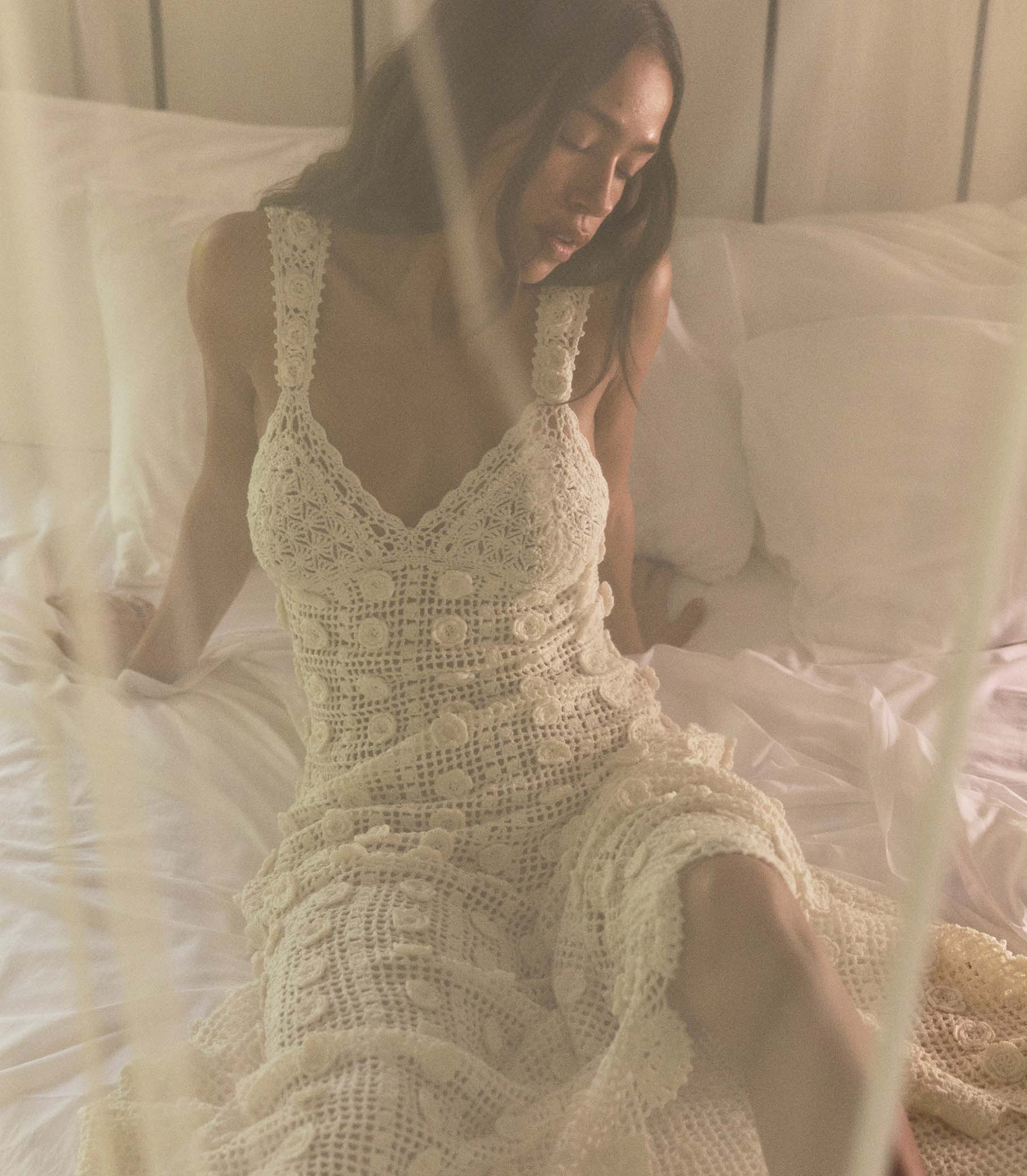 Woman wearing a white crochet dress sitting on a bed. 1
