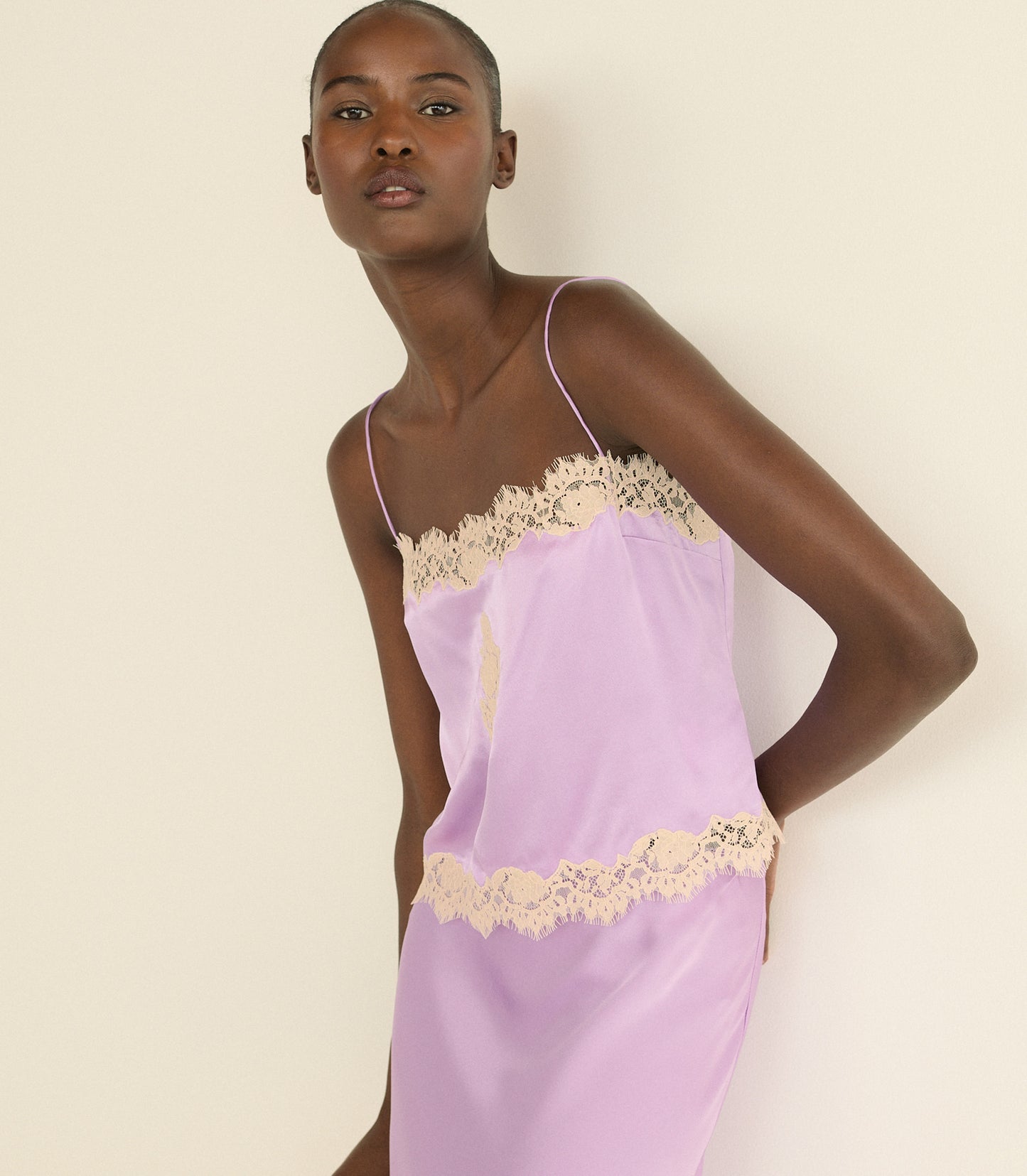 A model wearing a lavender satin top with beige lace trim, photographed against a solid cream background.