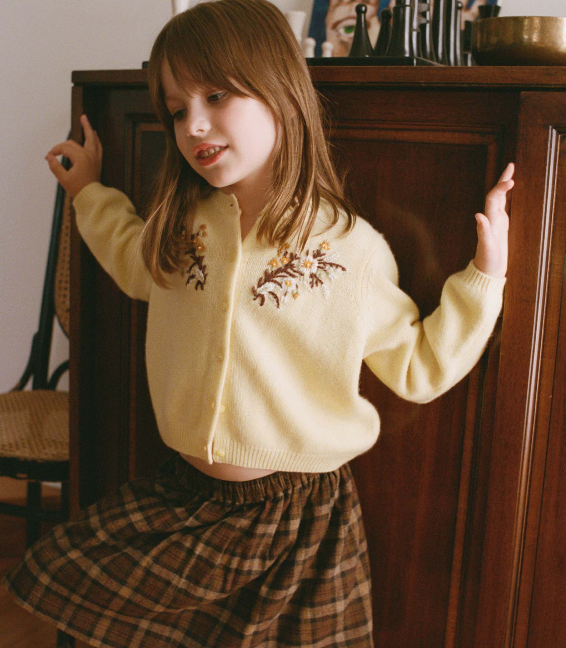 Young girl in a yellow cardigan with floral embroidery and plaid skirt standing in a room. 2
