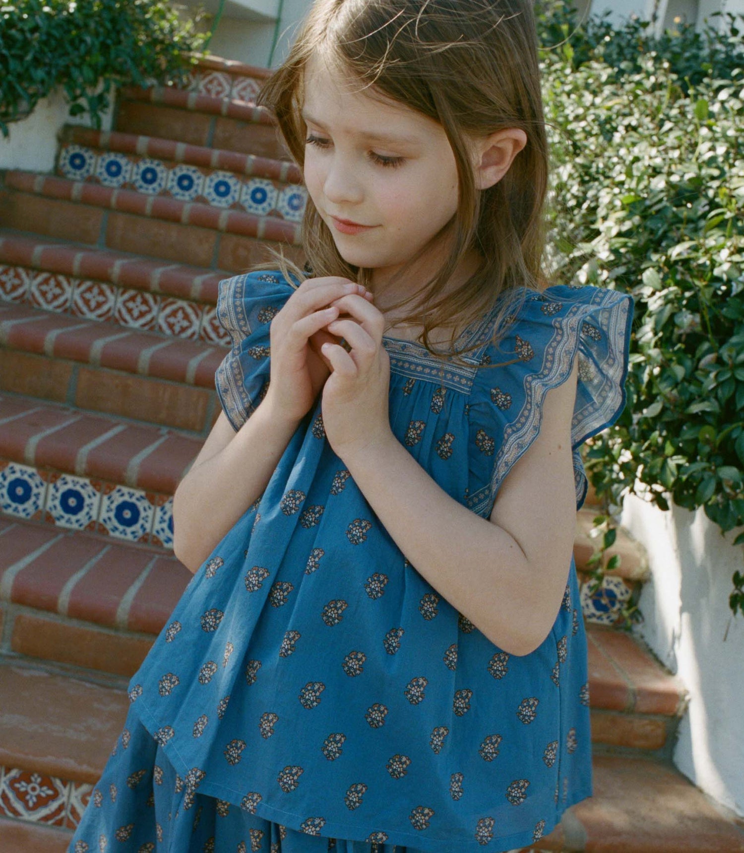 Young girl in a blue paisley top standing on outdoor steps with greenery.
