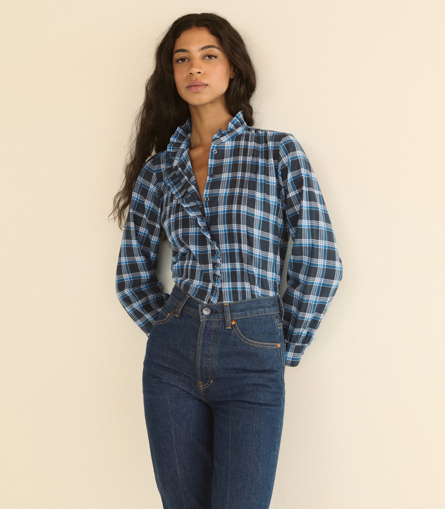 A woman wearing a blue and black plaid button-up shirt tucked into dark blue jeans, standing against a plain beige background.