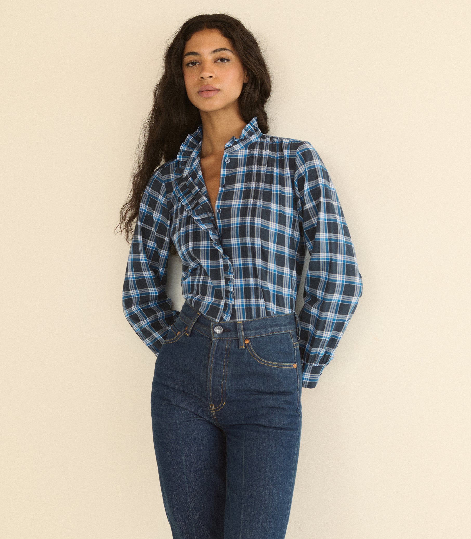 A woman wearing a blue and black plaid button-up shirt tucked into dark blue jeans, standing against a plain beige background. 1
