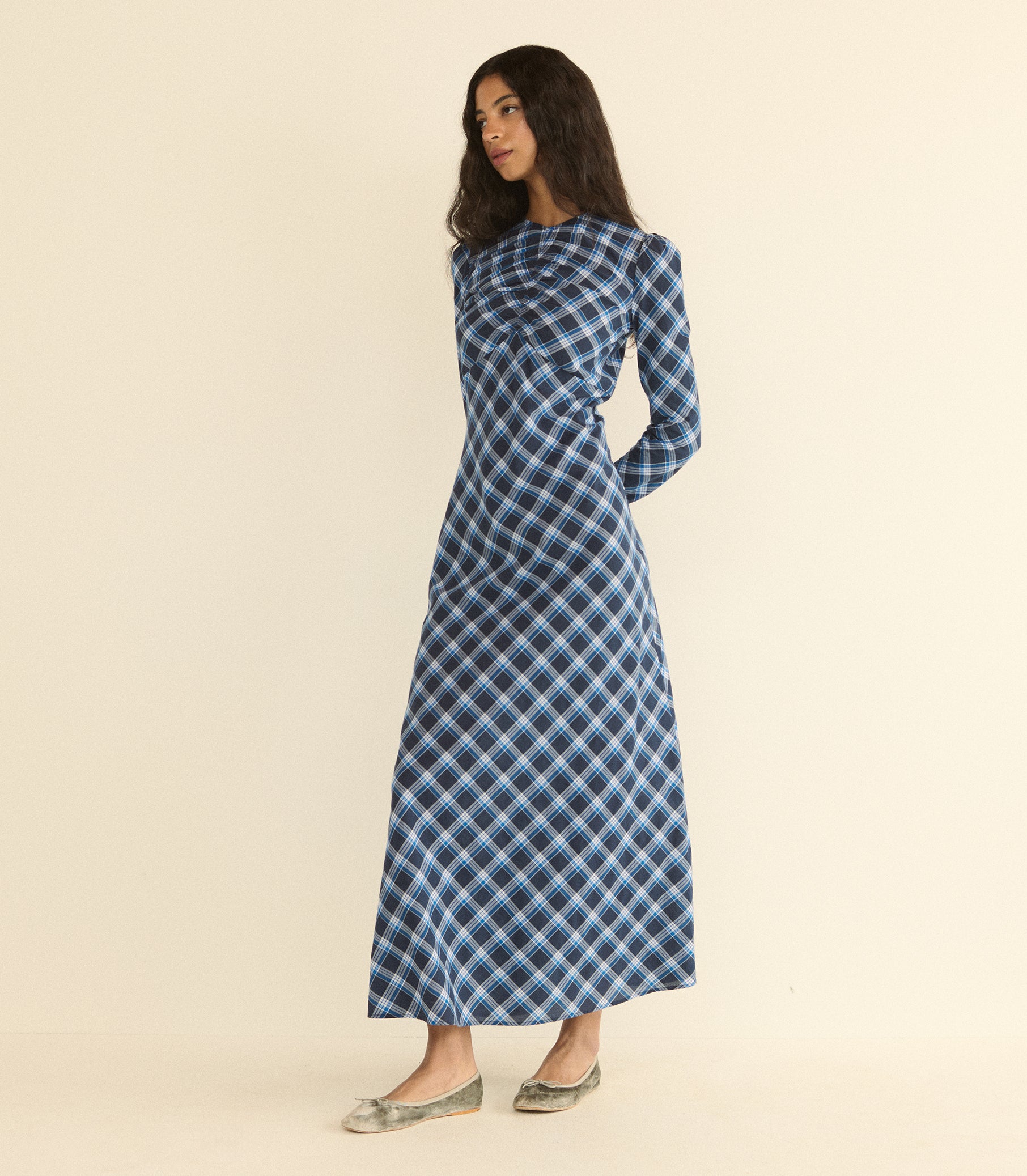 A woman modeling a long-sleeved, high-neck maxi dress with a blue, black, and white plaid pattern, standing against a solid beige studio background. 1
