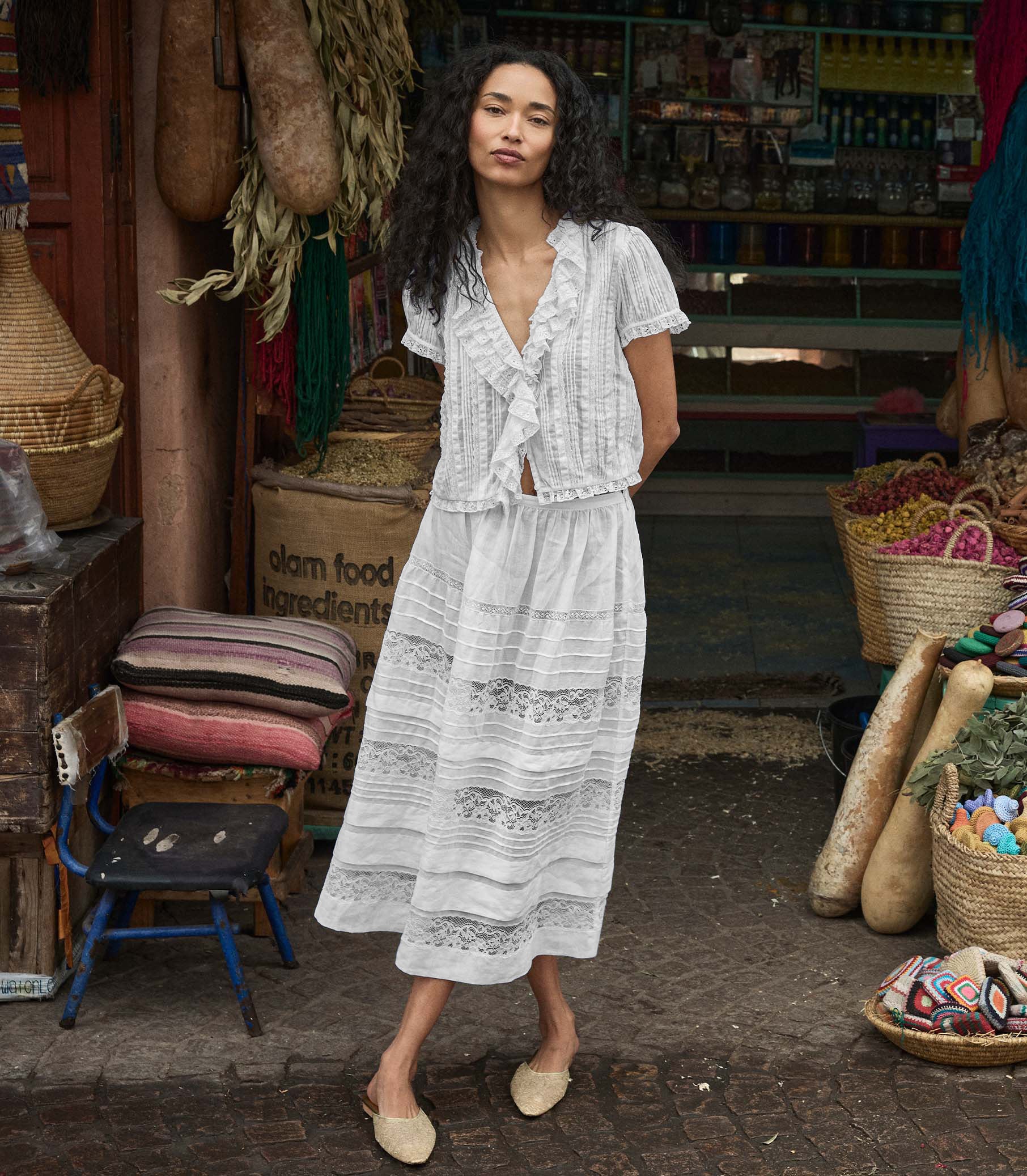 Woman in a white lace blouse and skirt standing in a market setting with various items around her.