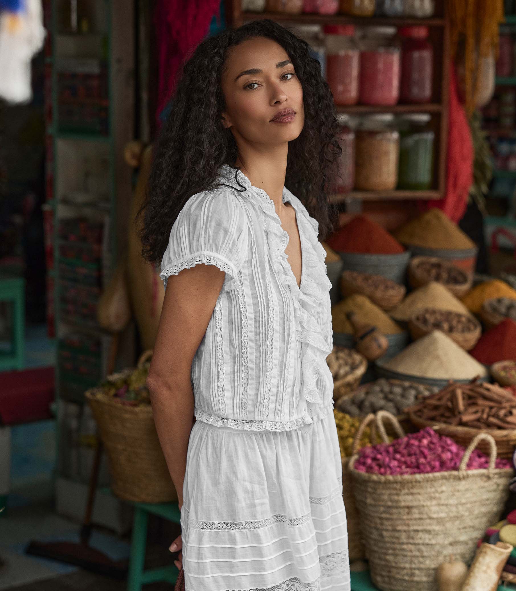 Woman in a white blouse standing in a market setting with various products and baskets. 1

