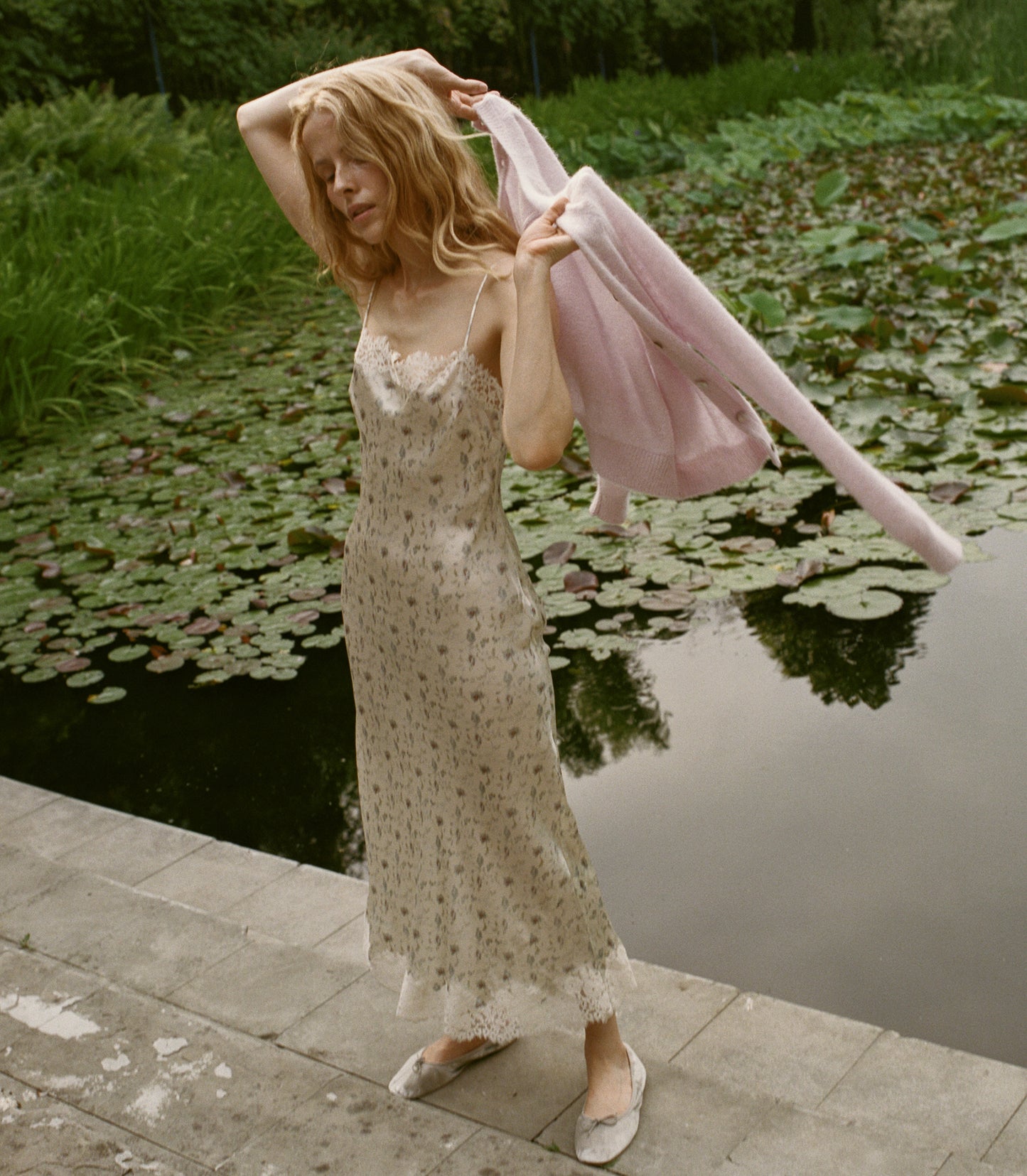 A woman stands on a stone walkway beside a pond with lily pads, wearing a light beige floral slip dress and holding a pale pink cardigan in front of ponds filled with lily pads.