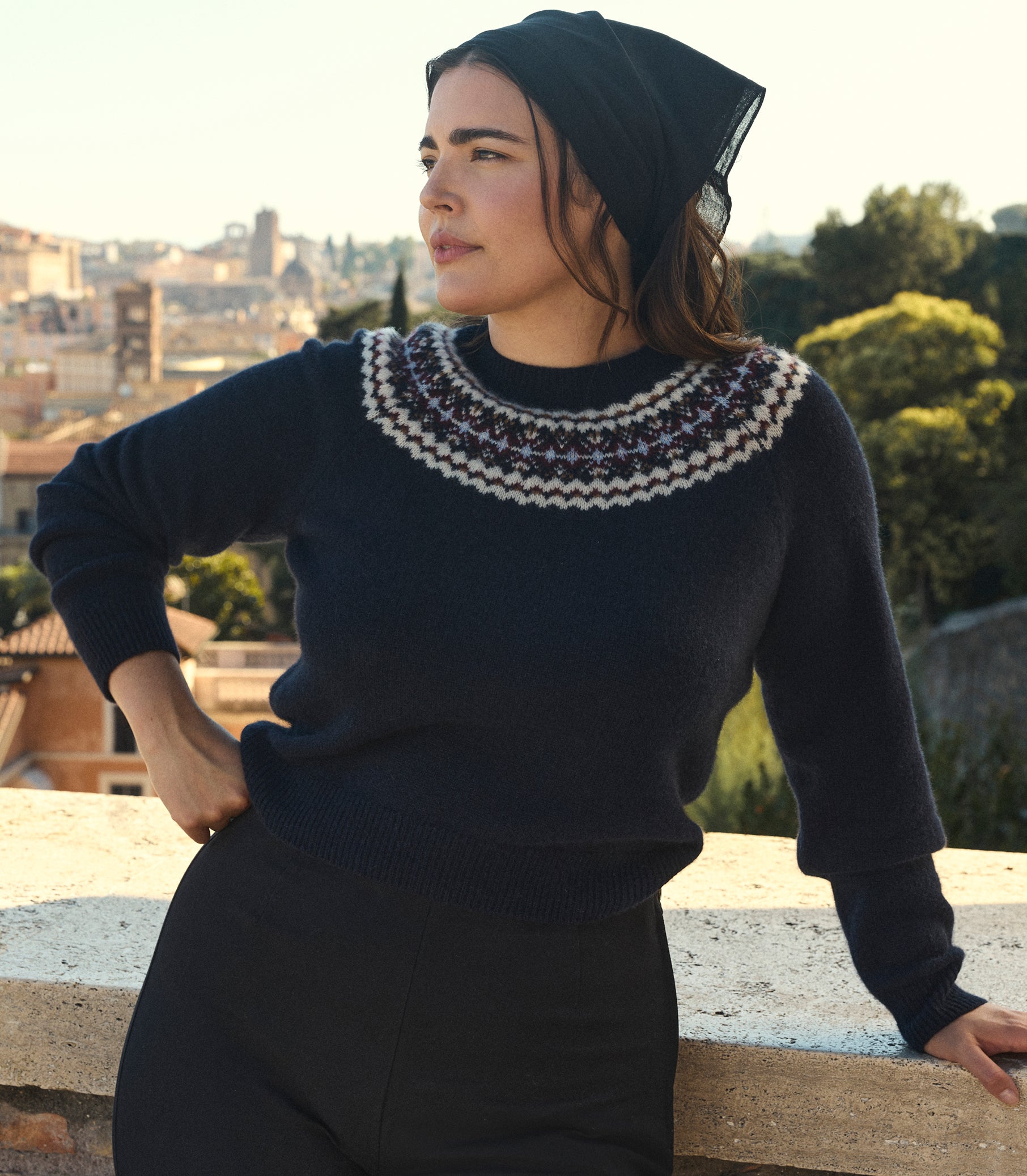 A woman wearing a dark navy knit sweater with a white and purple Fair Isle yoke pattern, paired with a black bandana, posing outdoors against a scenic city background. 1
