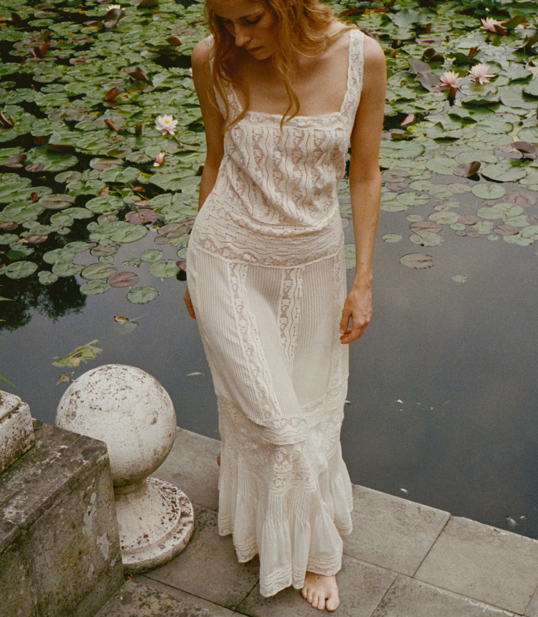 A woman wearing a long cream lace dress stands barefoot on a stone ledge beside a pond covered with lily pads. 1

