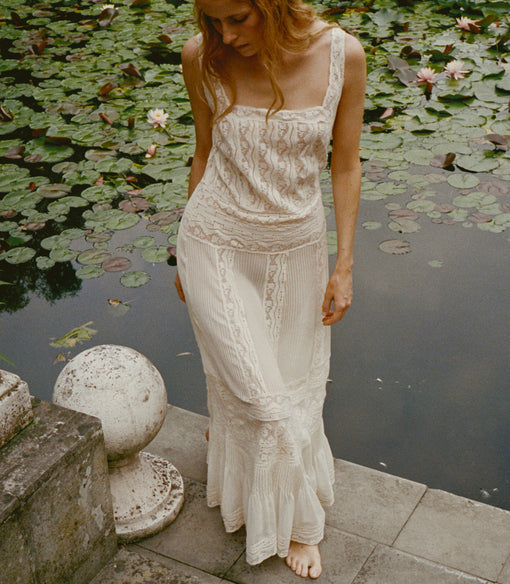 A woman wearing a long cream lace dress stands barefoot on a stone ledge beside a pond covered with lily pads.