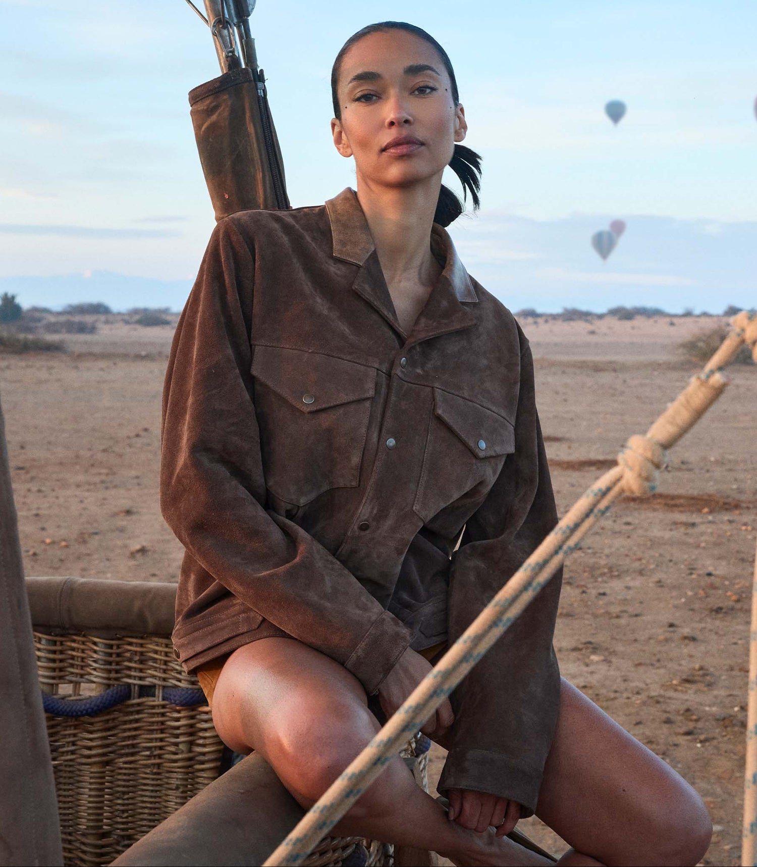 Person in a brown jacket sitting on a hot air balloon basket with balloons in the background