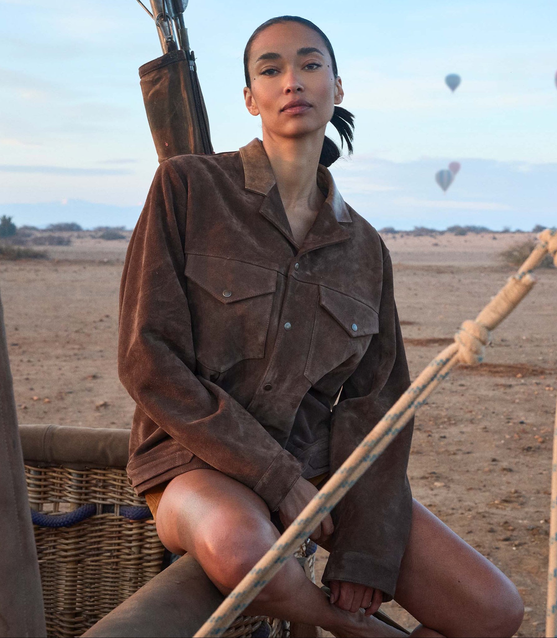 Person in a brown jacket sitting on a hot air balloon basket with balloons in the background