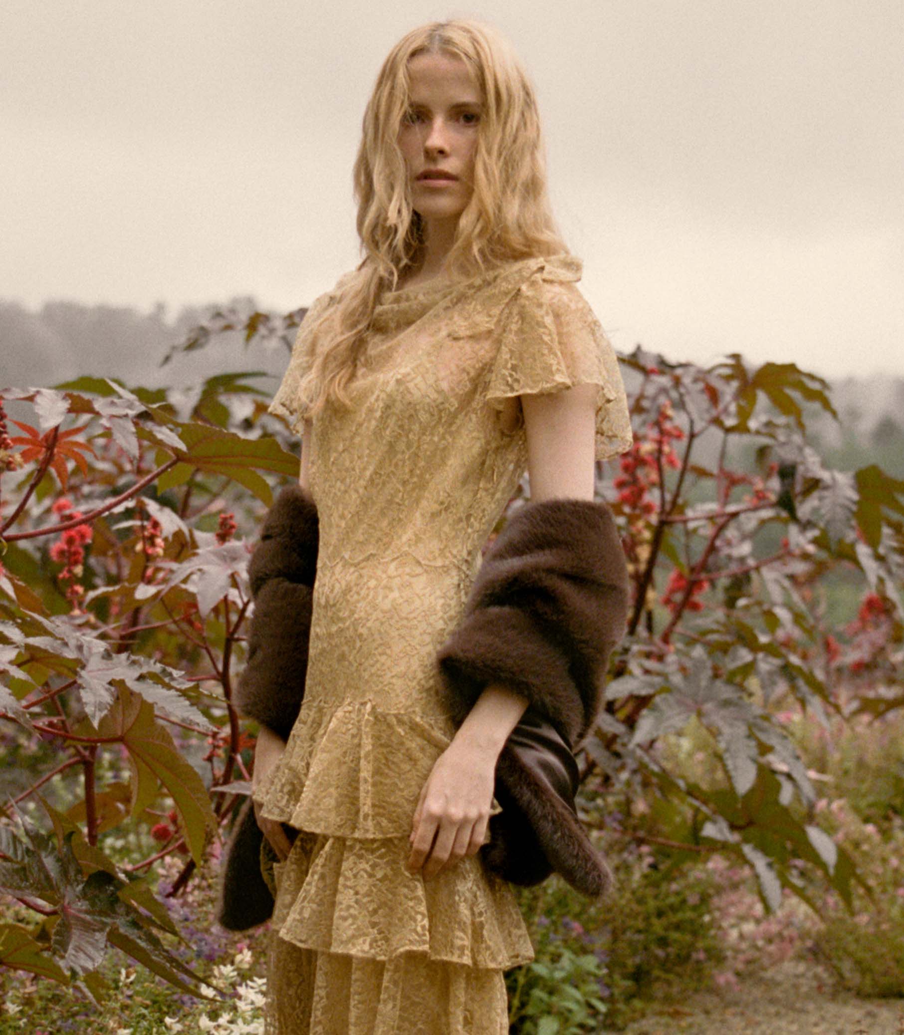 Woman in a gold lace dress holding a faux fur stole amidst autumn foliage.