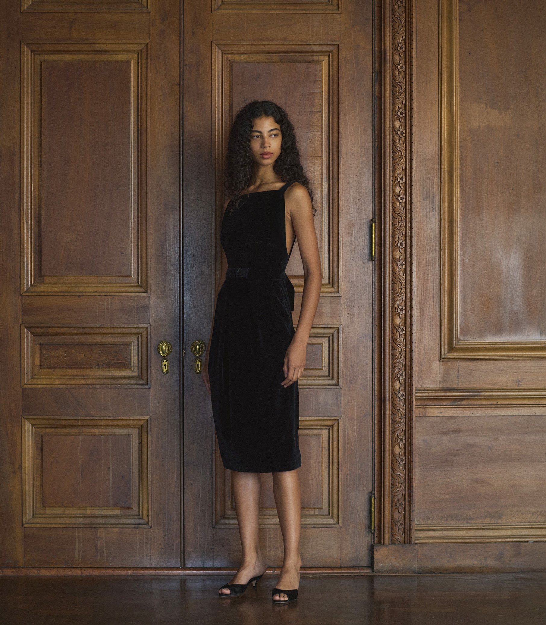 Woman in a black velvet dress standing in front of a large wooden door.