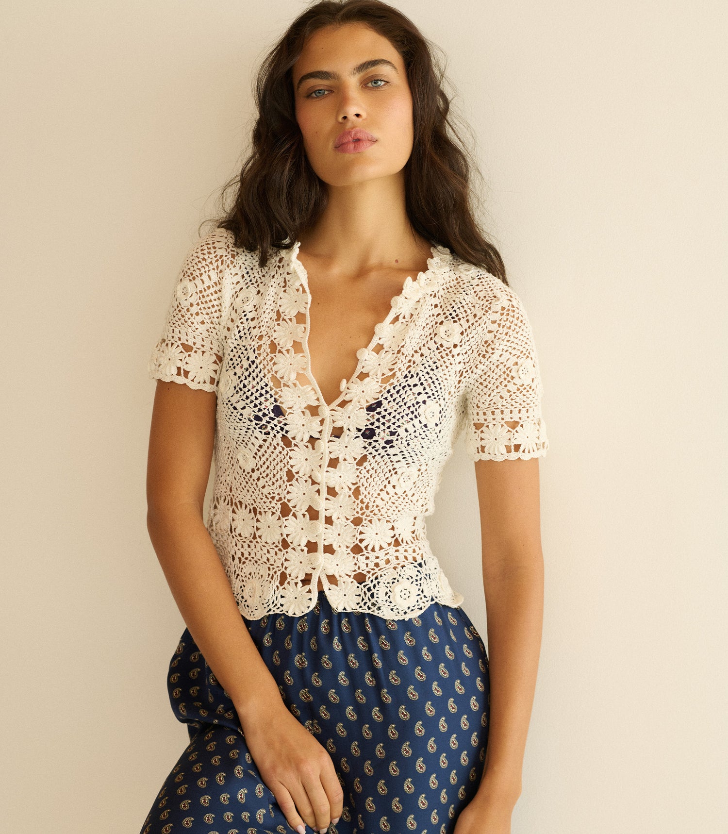 Woman wearing a white crochet top and blue patterned pants against a beige background