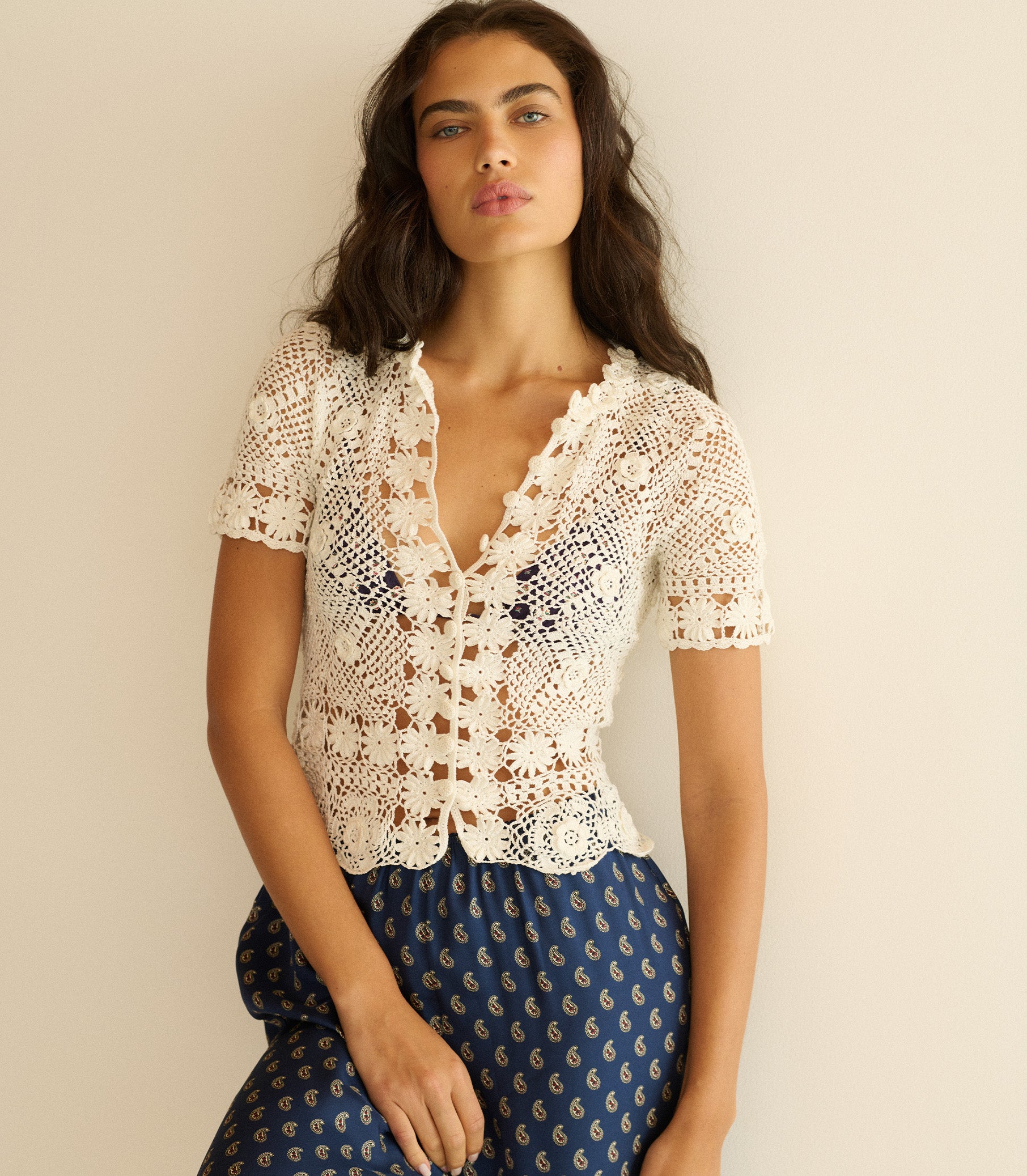 Woman wearing a white crochet top and blue patterned pants against a beige background 1
