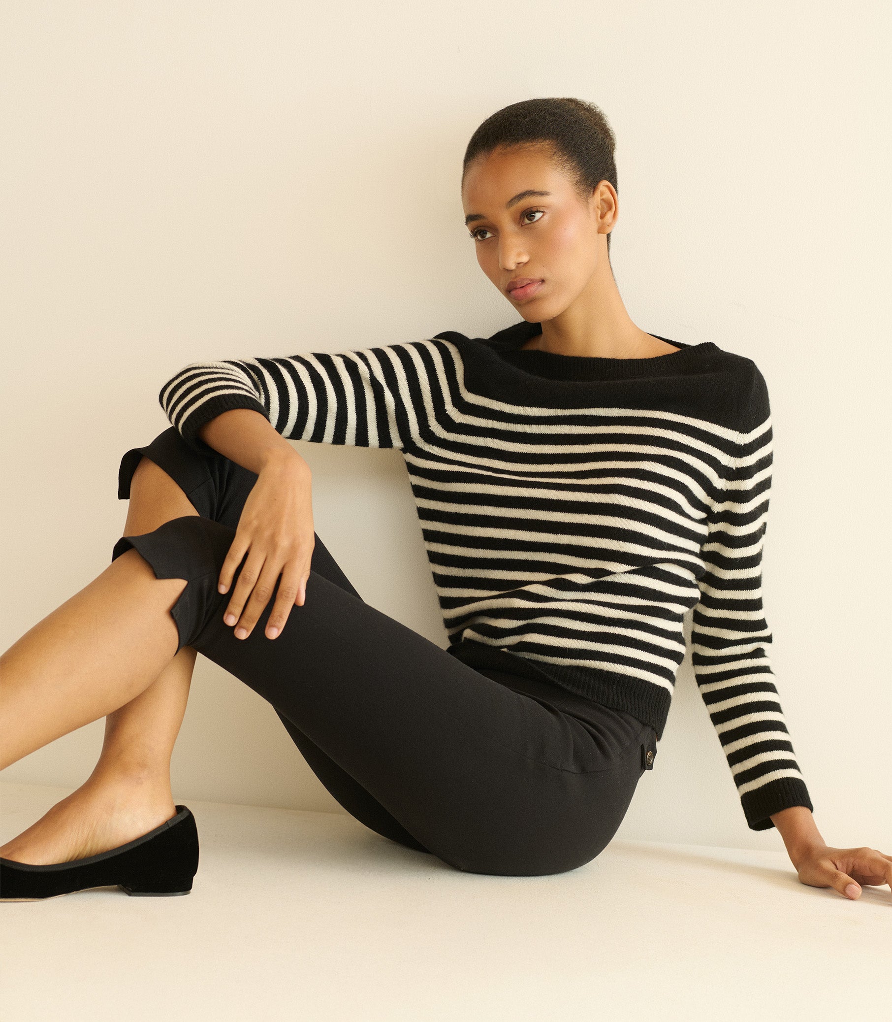 Woman wearing a black and white striped sweater and black pants sitting against a beige wall.