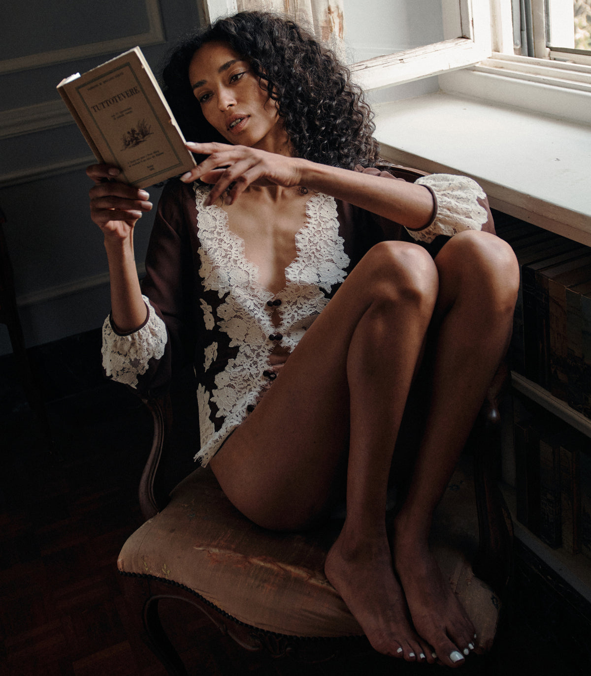 A woman with curly dark hair sits barefoot on a cushioned chair by a window, holding and reading a book. She is wearing a sheer white lace lingerie-style top with a dark underlayer. 1
