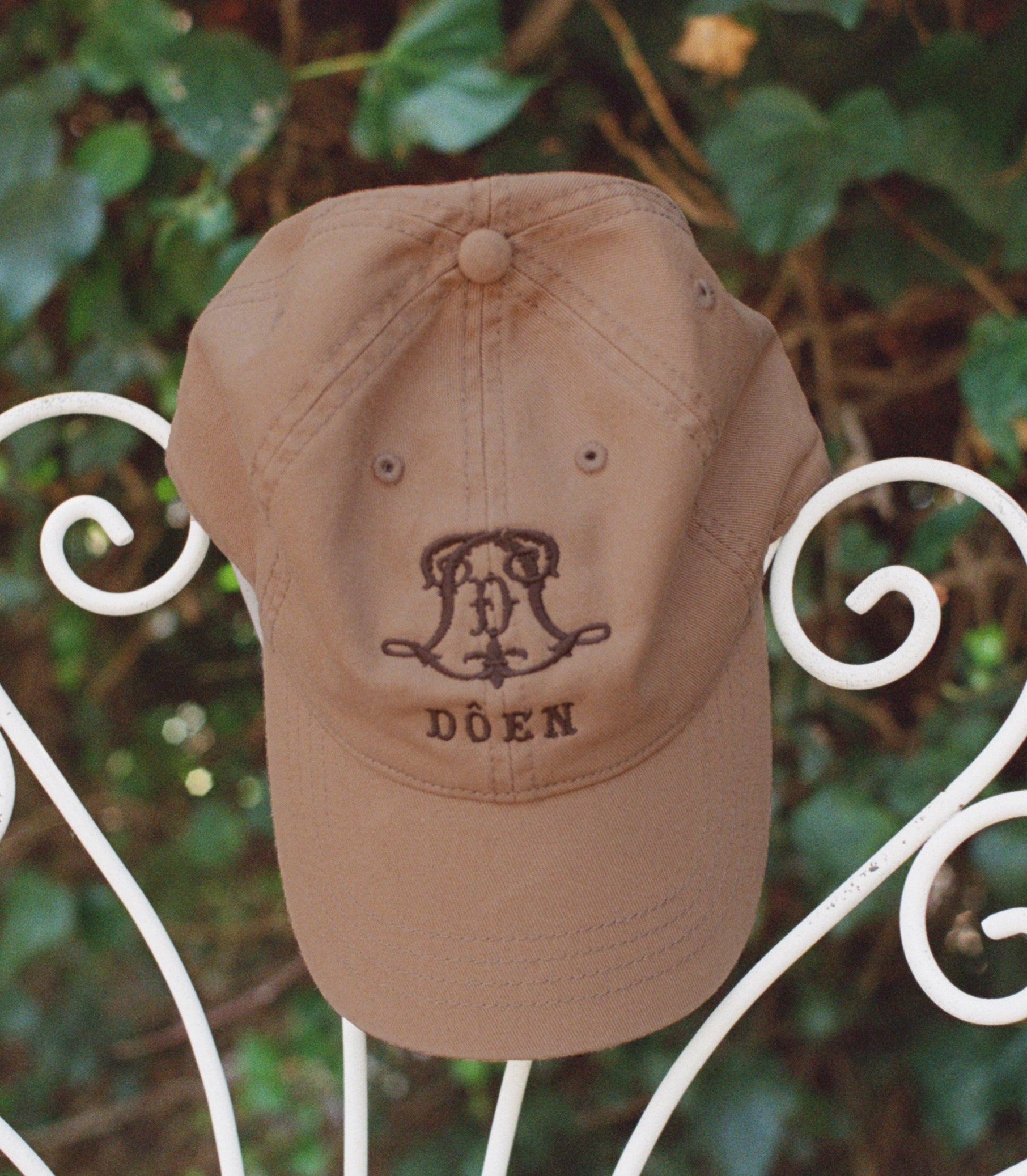A tan baseball cap with a dark brown embroidered monogram and the word 'DÔEN' on the front, displayed on a white wrought iron garden chair with green foliage in the background.