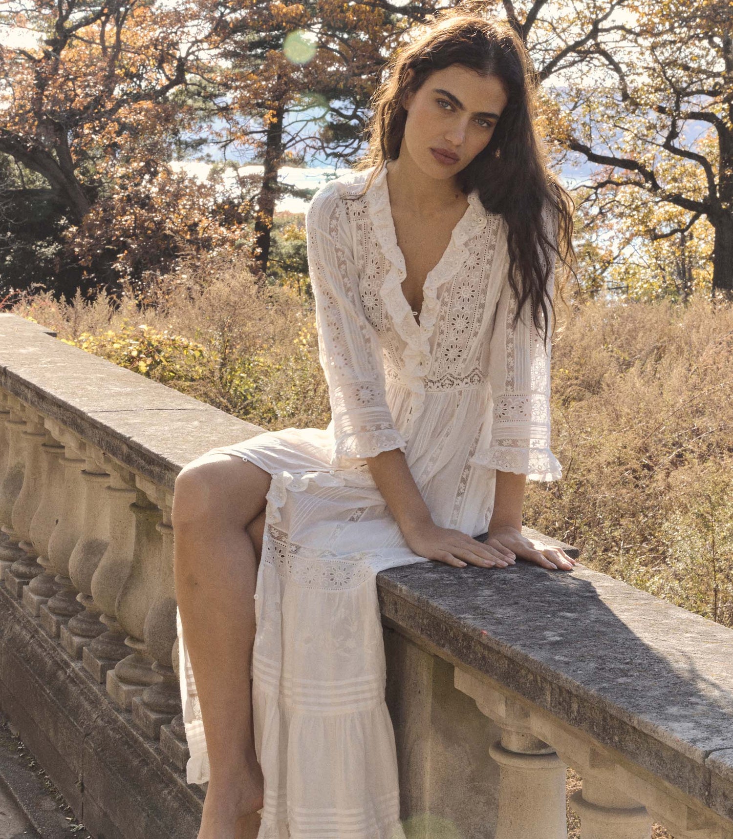 Woman in a white lace dress sitting on a stone wall with trees and grass in the background