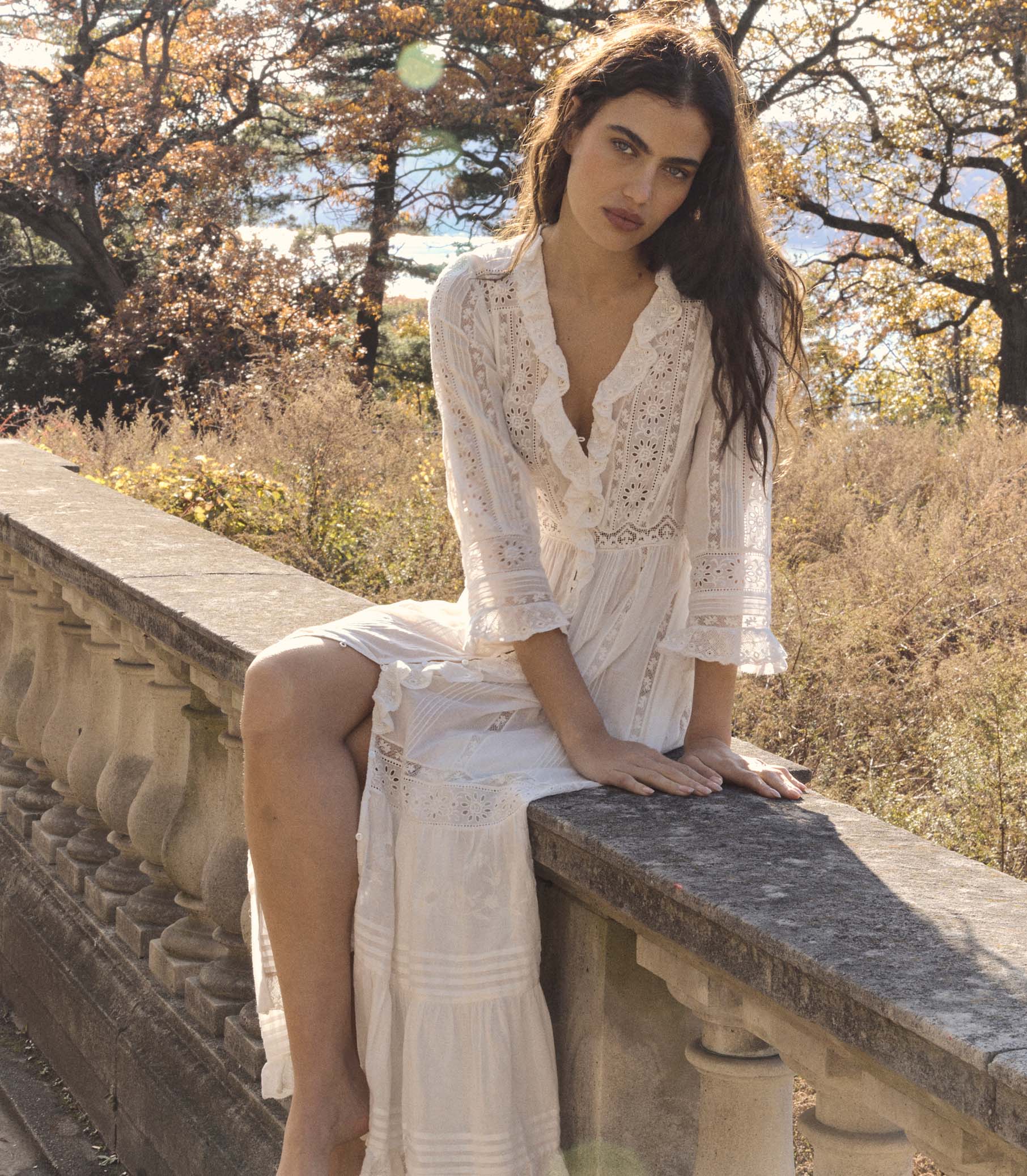 Woman in a white lace dress sitting on a stone wall with trees and grass in the background 1
