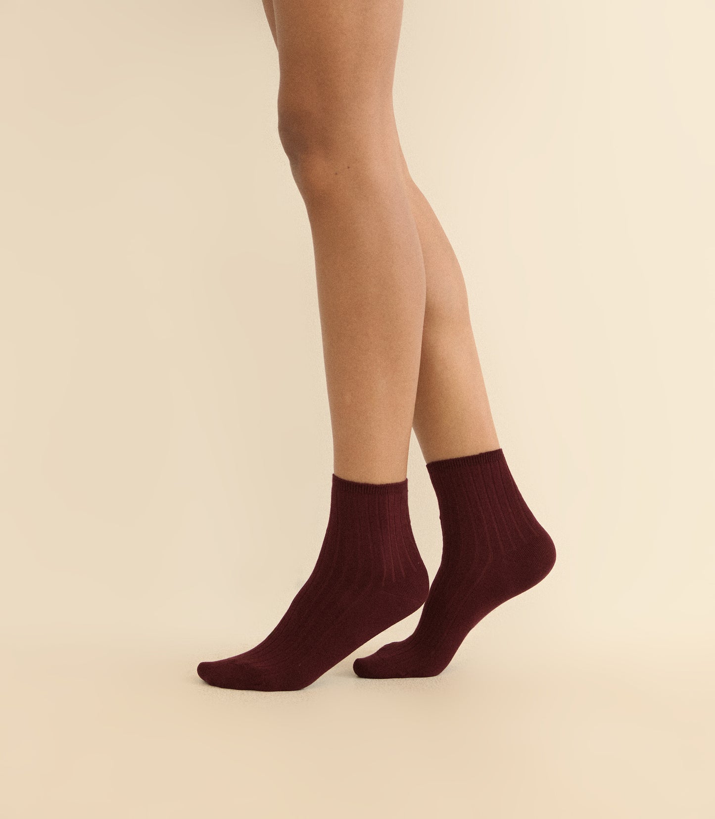 A pair of ankle-length ribbed socks in deep burgundy, worn on a model's feet against a solid light beige background.