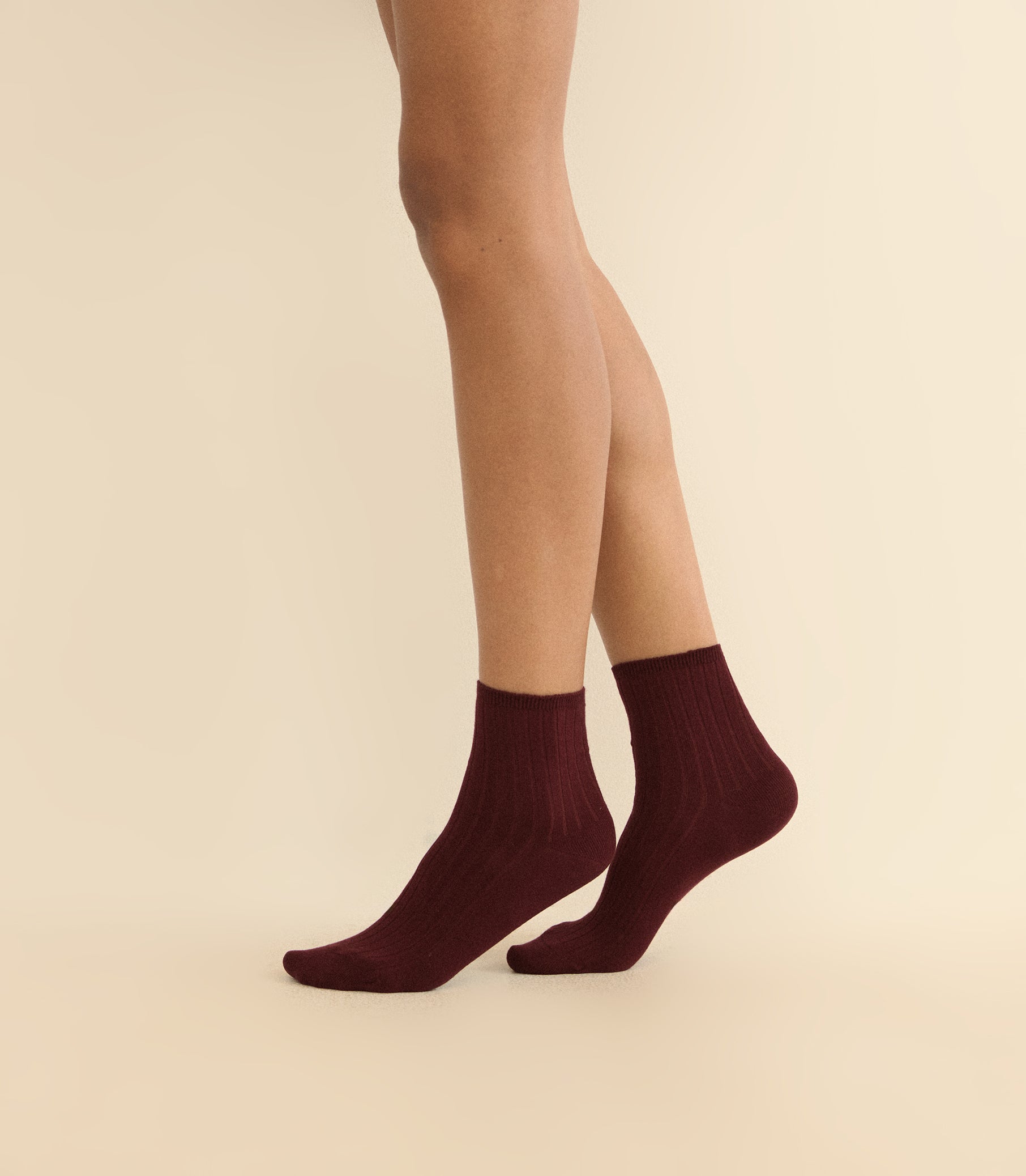 A pair of ankle-length ribbed socks in deep burgundy, worn on a model's feet against a solid light beige background. 1
