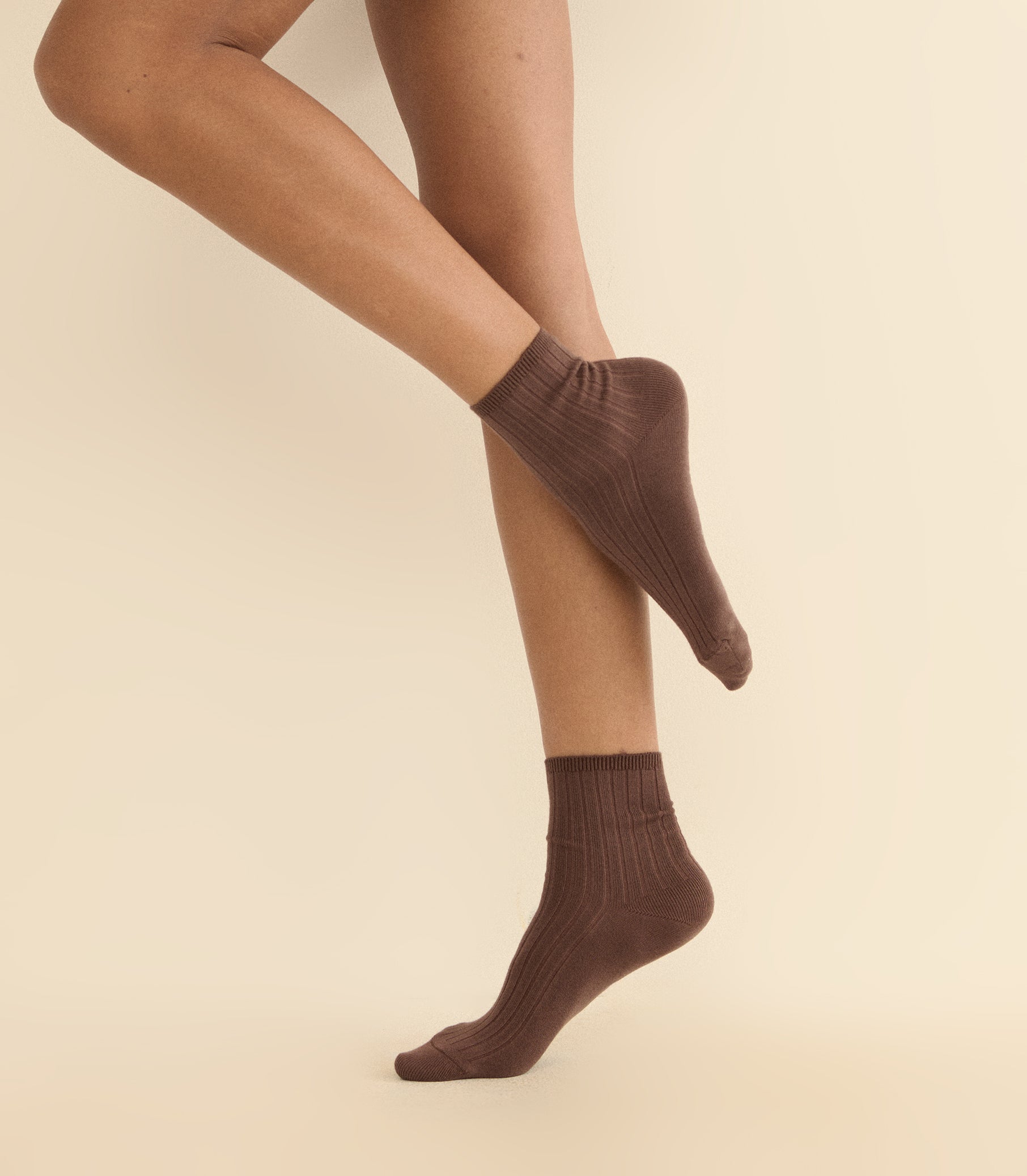 A person wearing ribbed ankle socks in a dark brown color, shown from the knees down against a plain beige studio background.