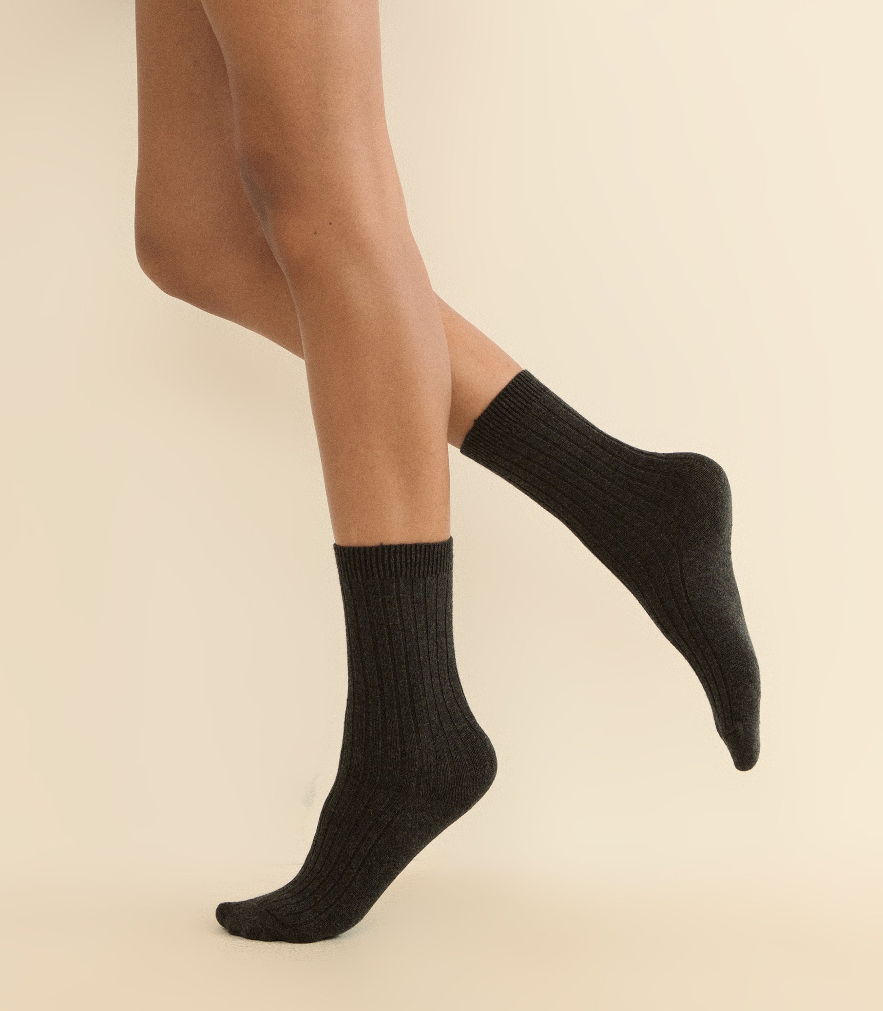 A pair of dark charcoal gray ribbed ankle socks worn on a model's feet, shown against a solid light beige background. 1
